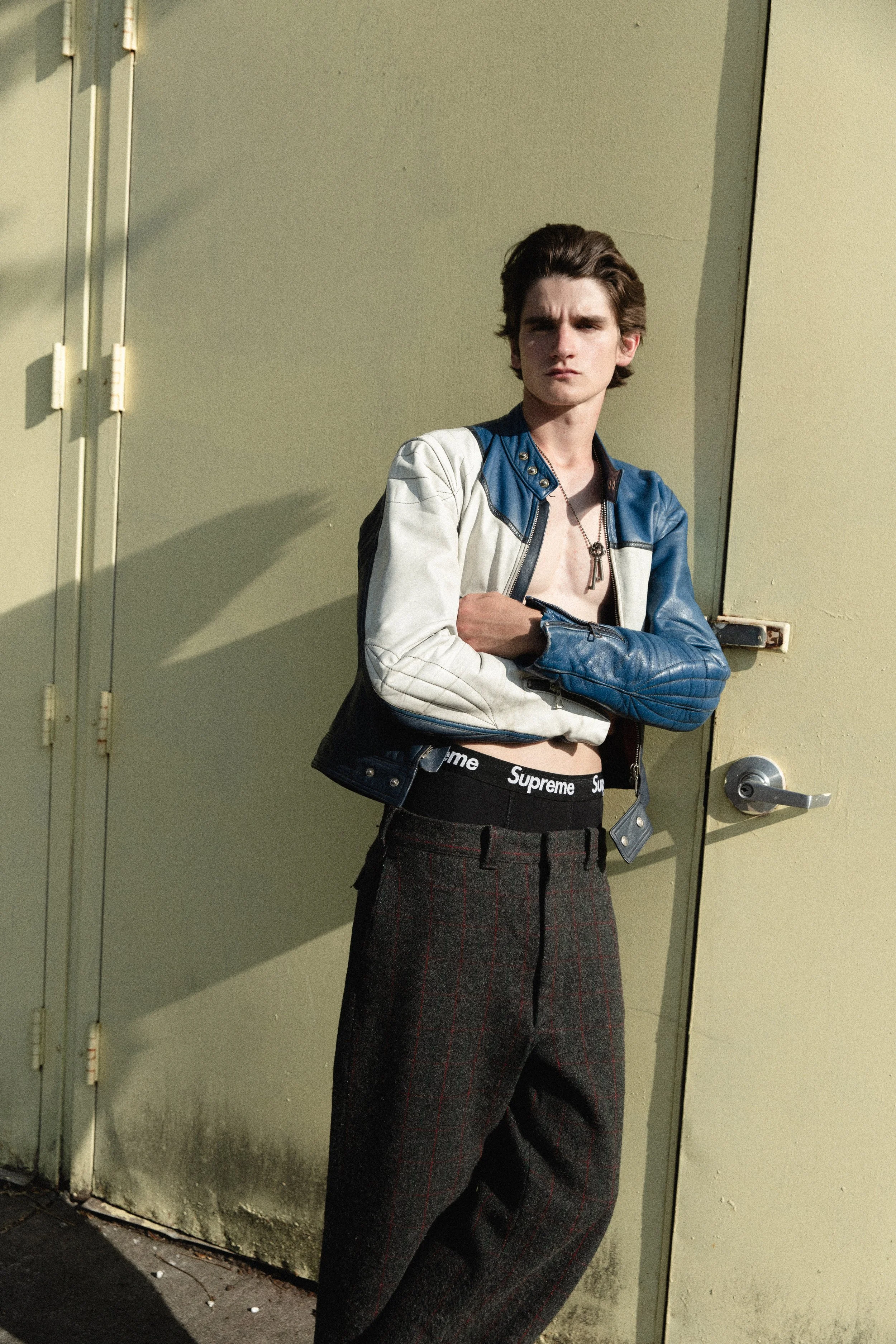 Young man standing against a beige wall with a door, wearing a color-blocked blue and white leather jacket, a black Supreme waistband, and loose dark gray plaid pants.