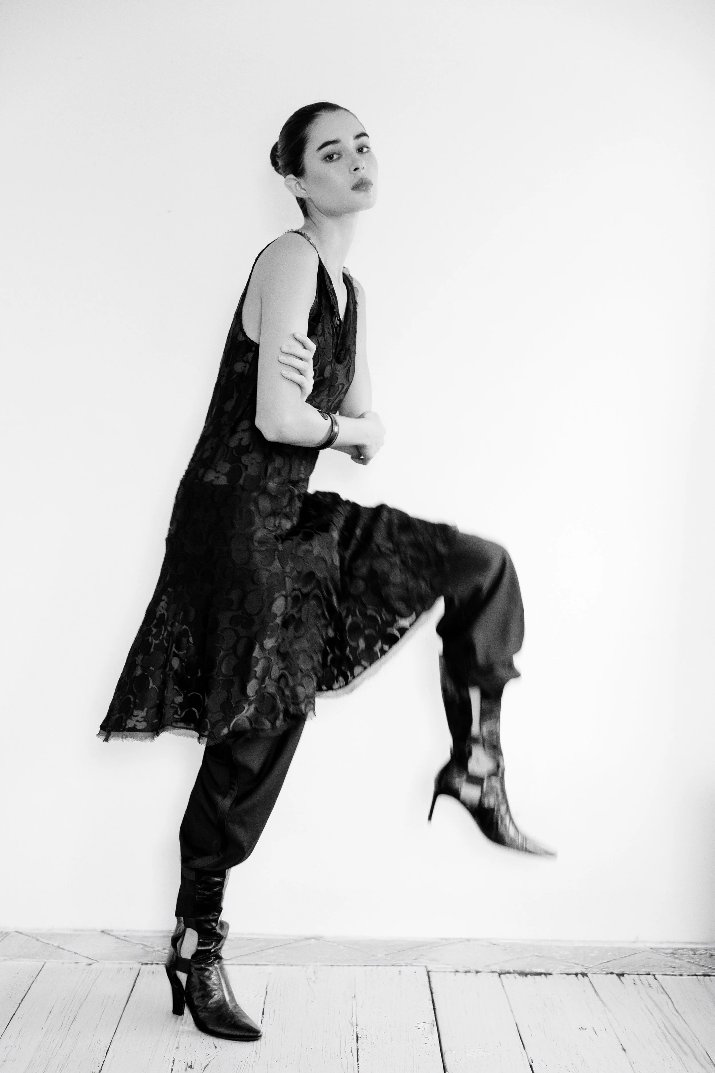 Black and white photo of a woman in high fashion, wearing a sleeveless, patterned dress and high-heeled boots, posed against a plain white wall.