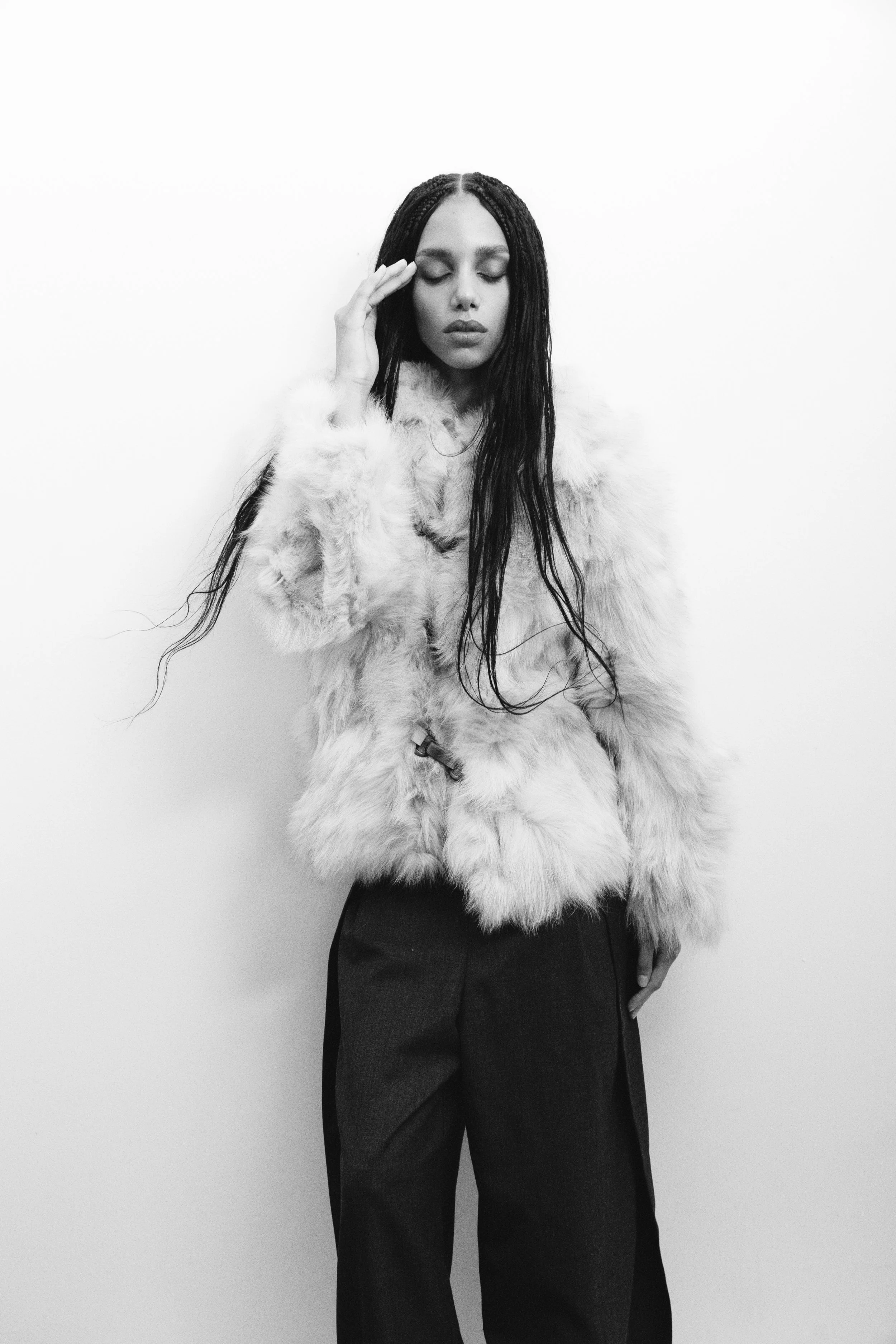 A woman with long dark hair wearing a fur coat and dark pants, standing against a plain white background, with her eyes closed and her hand touching her face.