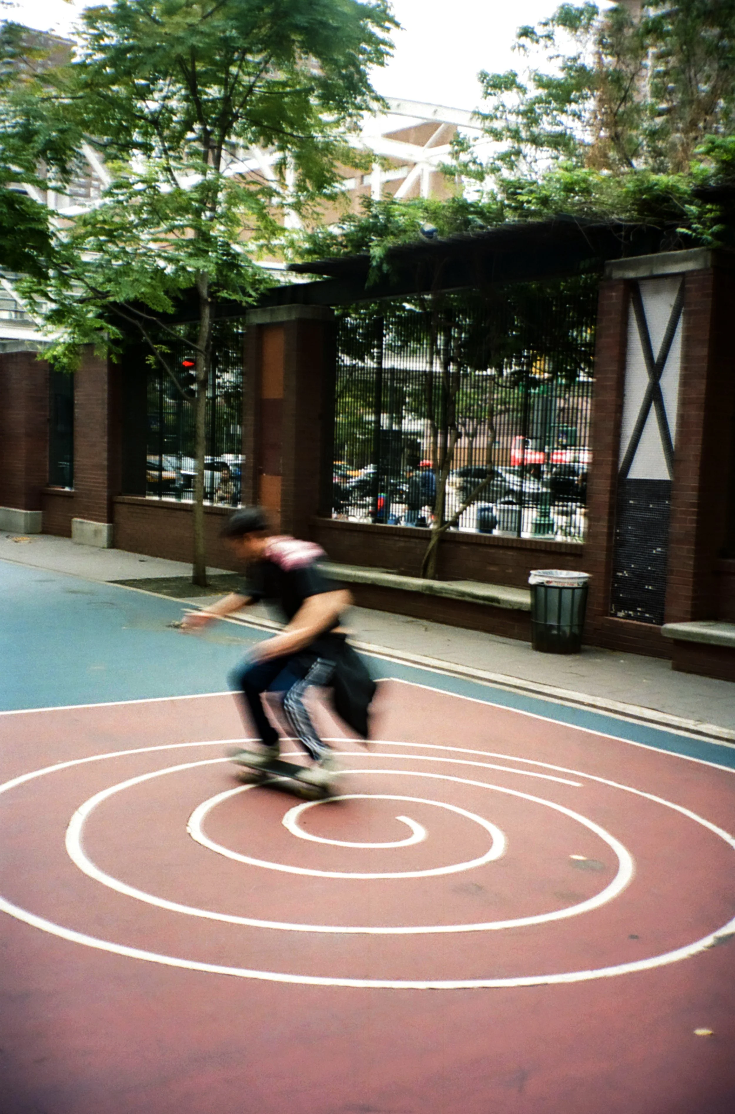 A person skateboarding on a spiral marking on an outdoor court, with trees and a brick building in the background.