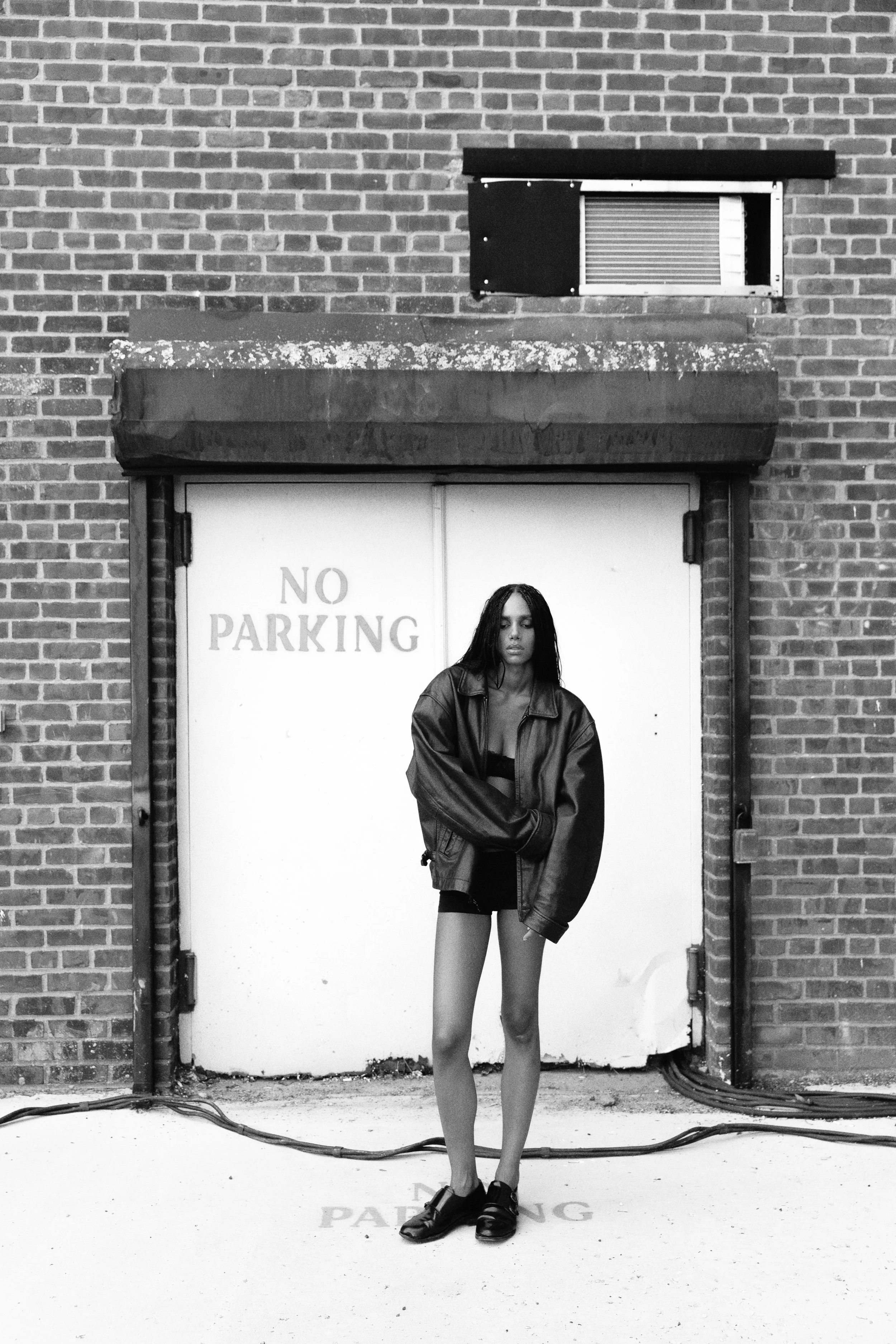 Black and white photo of a woman standing in front of a closed door with a "No Parking" sign on it. She has long dark hair, wears a leather jacket over a top, and has one leg slightly bent.