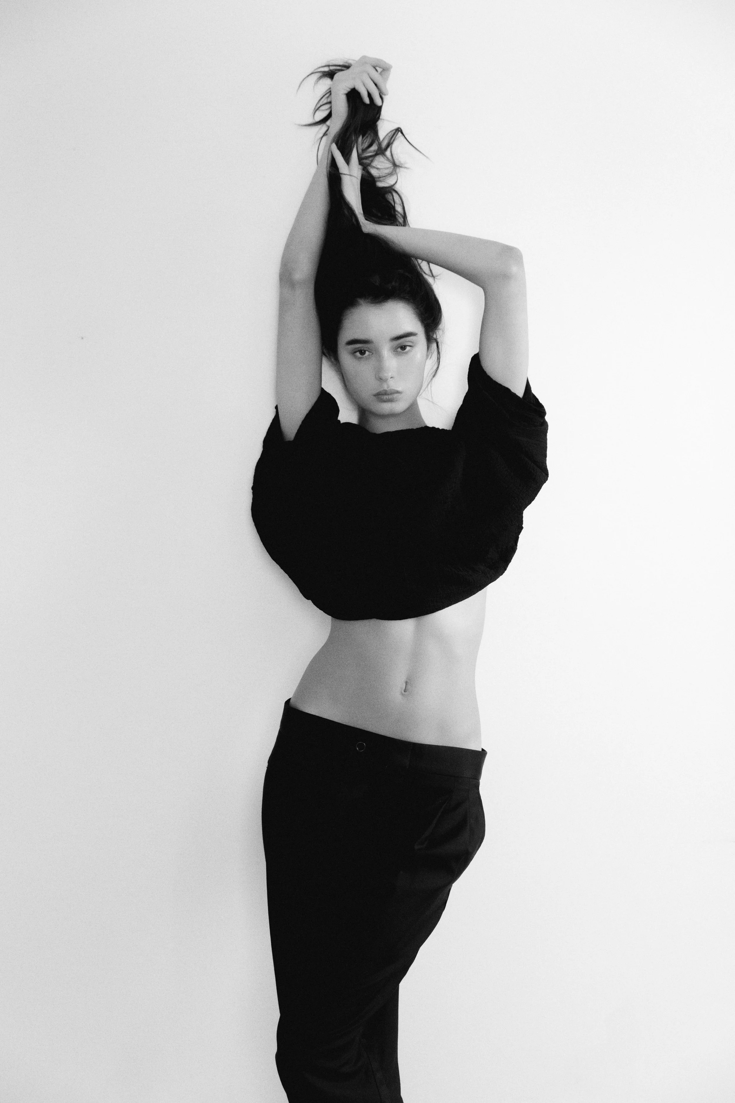 Black-and-white photo of a young woman with long hair, wearing a black shirt and black pants, striking a pose with arms raised and one leg slightly bent, against a plain white background.