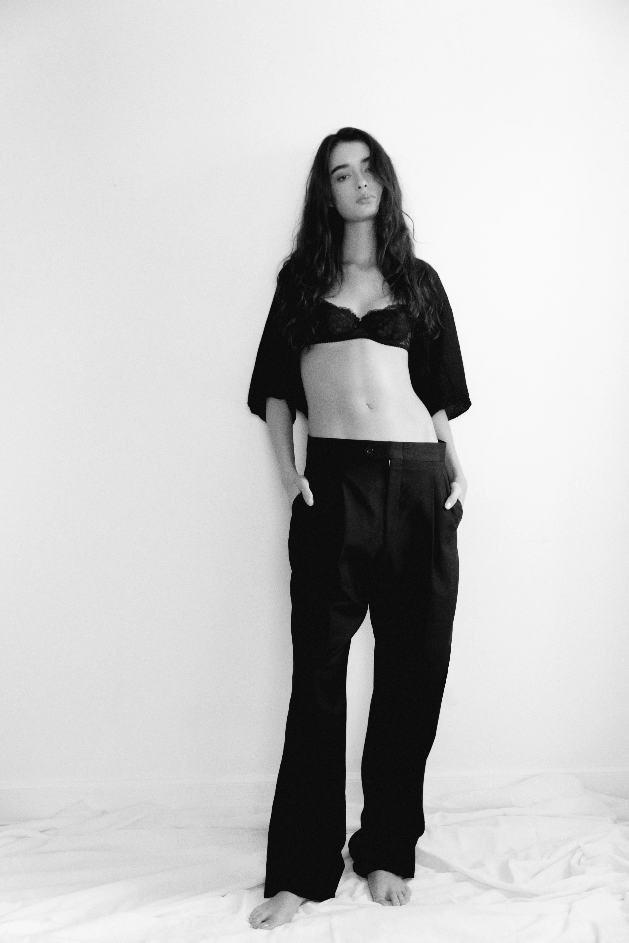 A young woman with long wavy hair standing against a plain wall. She is dressed in a black lace bra, cropped cardigan, and loose black pants, barefoot on a white sheet on the floor.