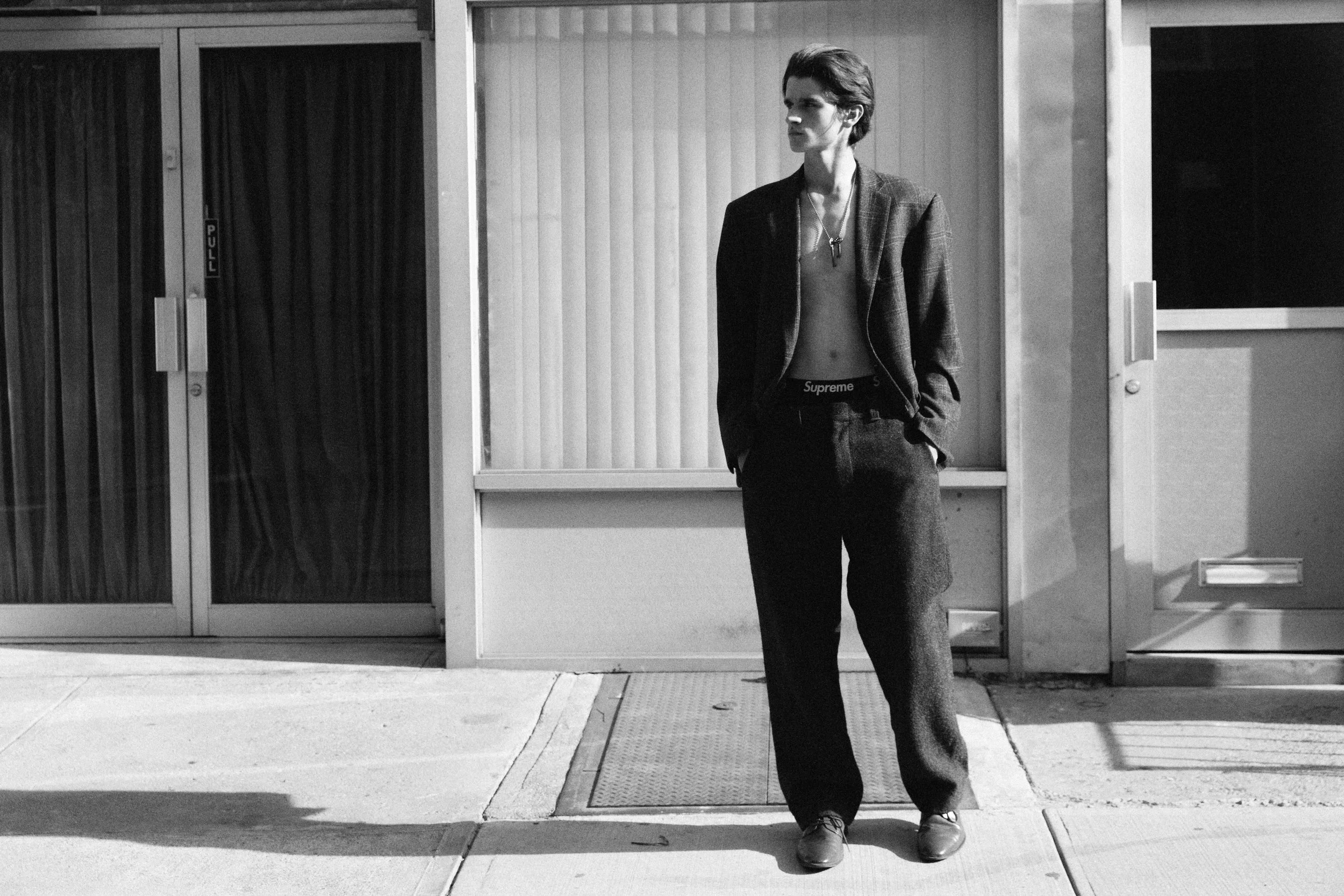A young man with dark hair, dressed in a blazer, shirt, and loose pants, stands outside near a glass door with vertical blinds on the window, illuminated by sunlight, in black and white.