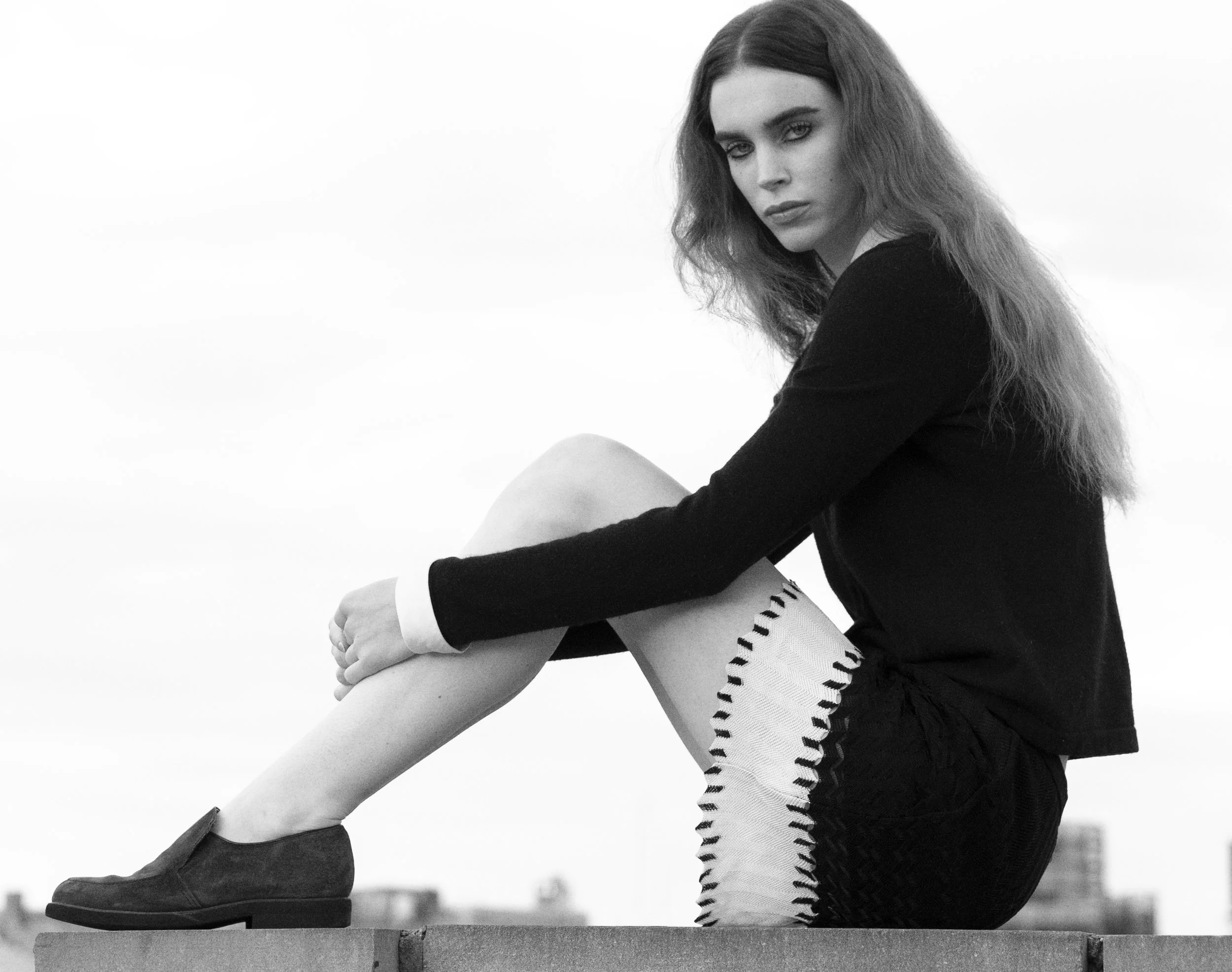Black and white photo of a woman sitting on a ledge with her arms wrapped around her knees, looking at the camera with a serious expression.