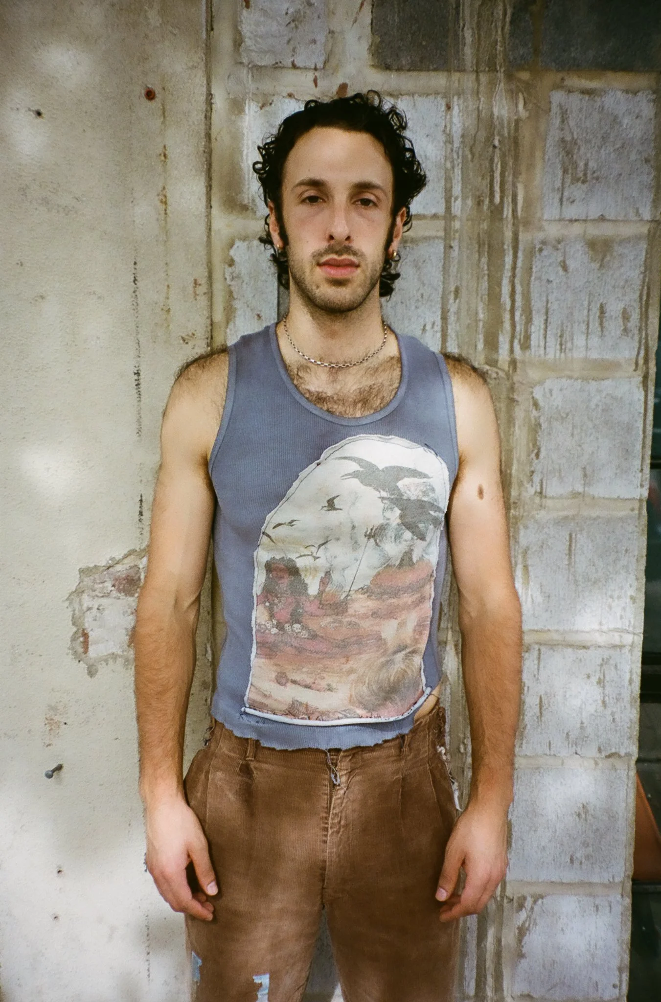 A young man with dark curly hair and light skin, wearing a sleeveless graphic tank top and brown pants, stands against a rustic concrete wall.