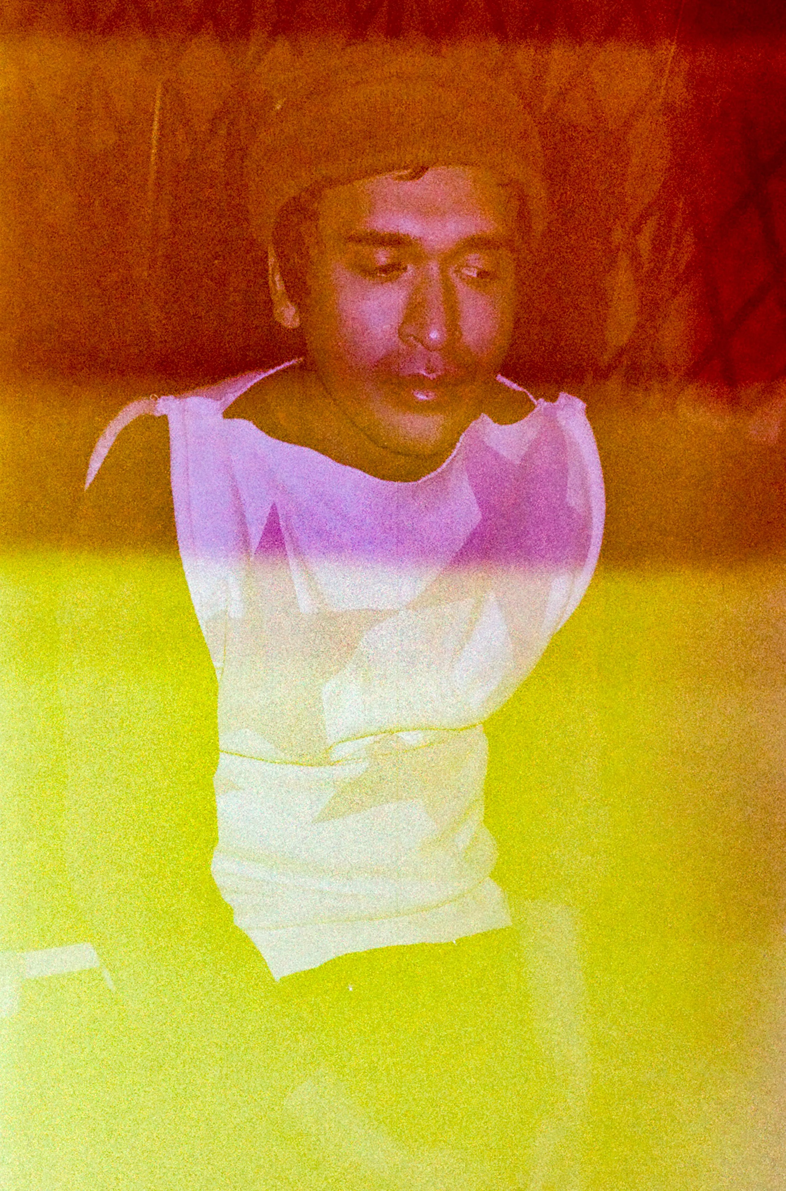 A young man with short dark hair and closed eyes, wearing a sleeveless white shirt, is sitting with a colorful, radiating background of red, yellow, and purple hues.