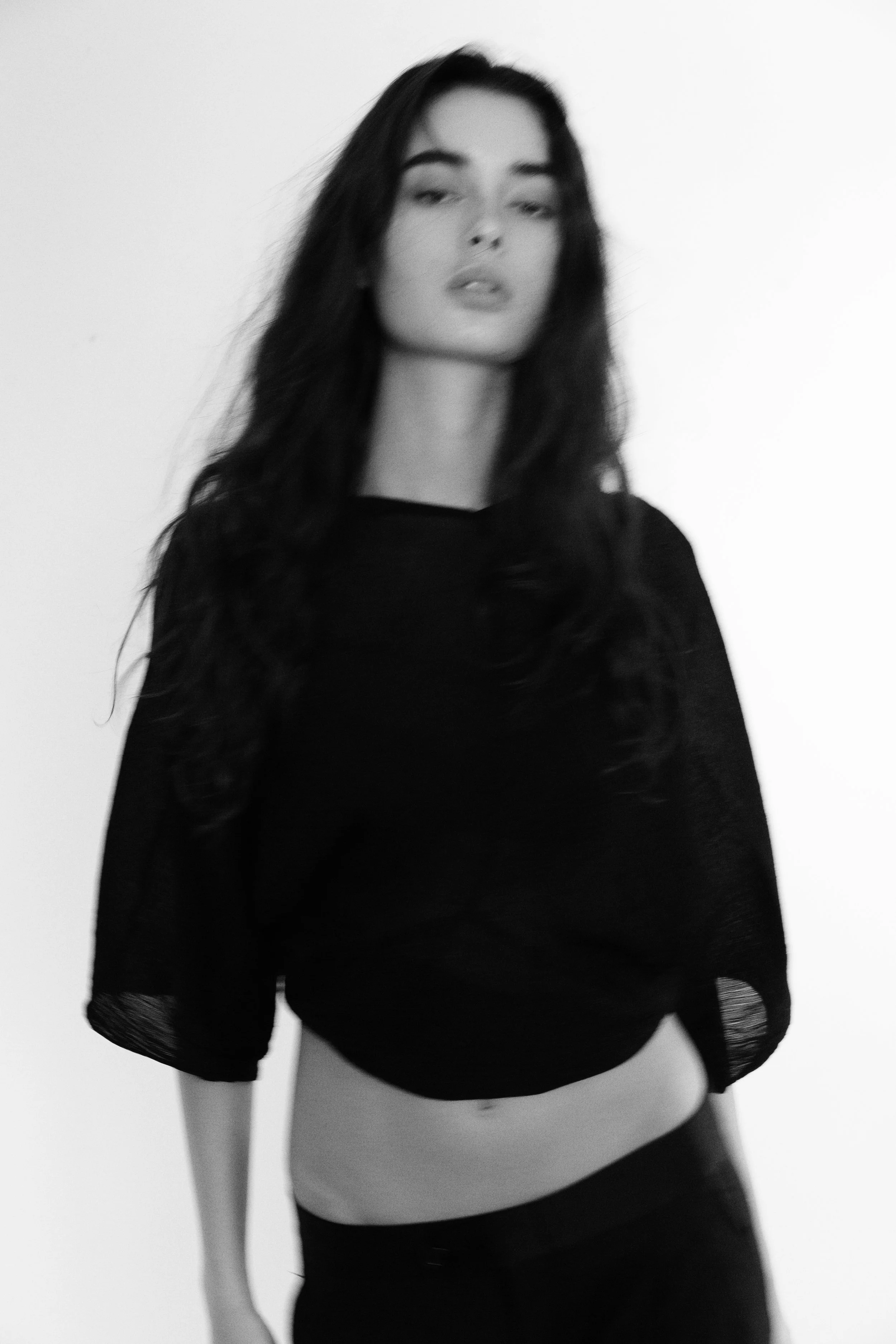 Black and white photo of a woman with long hair, wearing a black top, standing against a plain background.