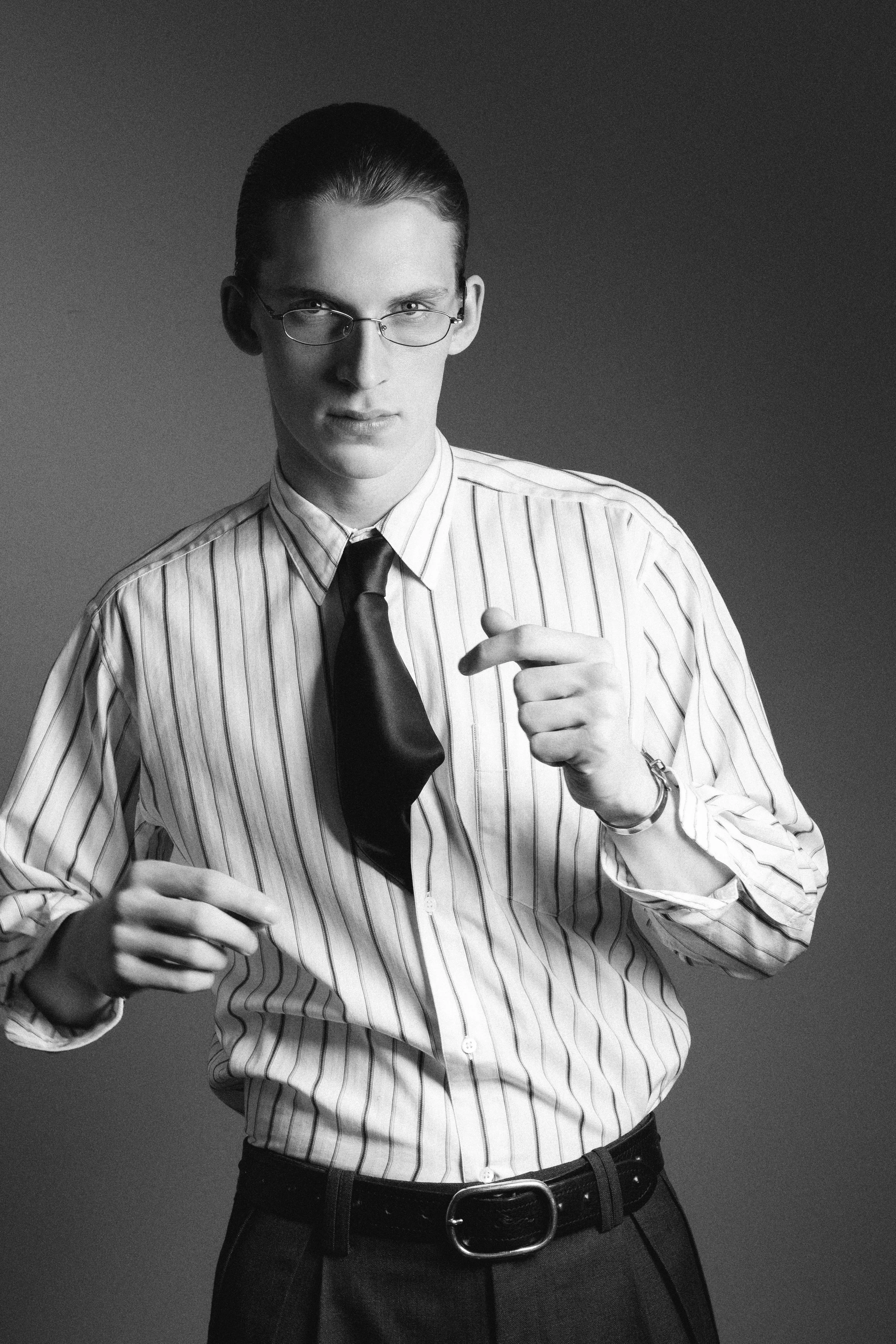 Black and white photo of a young man wearing glasses, a striped shirt, a black tie, and dark pants with a belt, standing against a plain background.