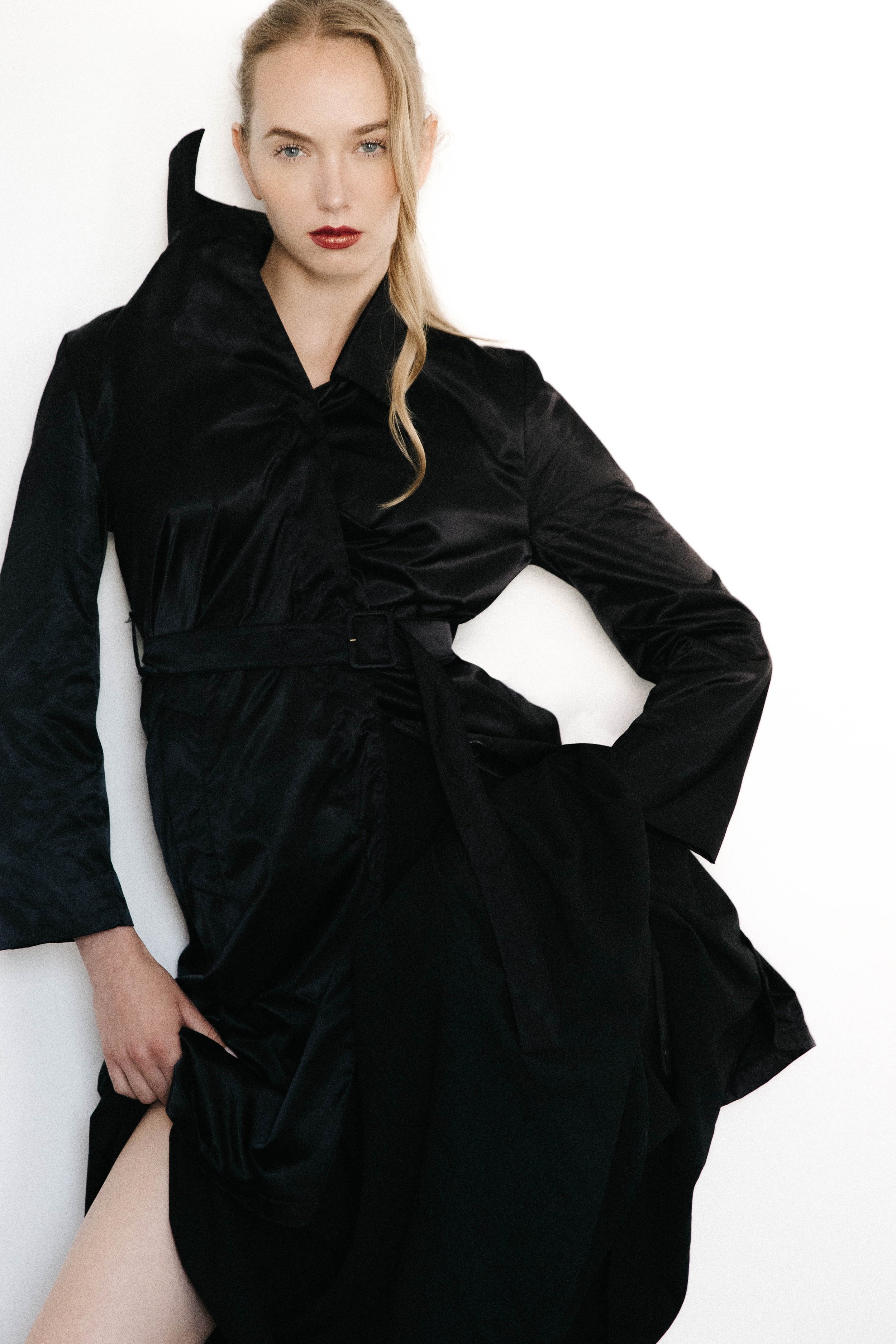 A young woman with blonde hair and red lipstick wearing a black satin trench coat, sitting against a white background.