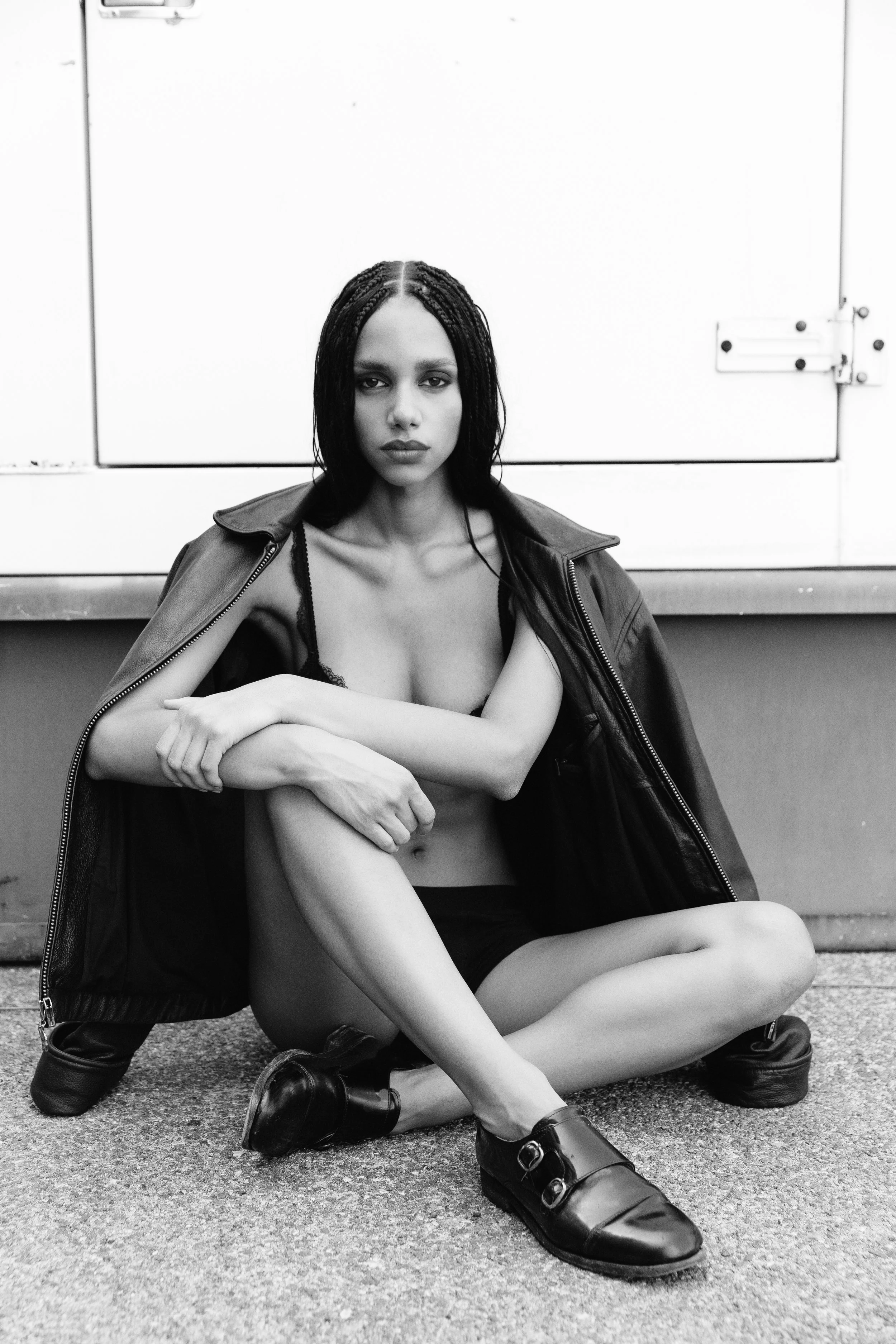 Black and white photo of a young woman with braided hair, sitting on the ground outside against a plain background, wearing a leather jacket and boots, with her legs crossed and arms resting on her knees.