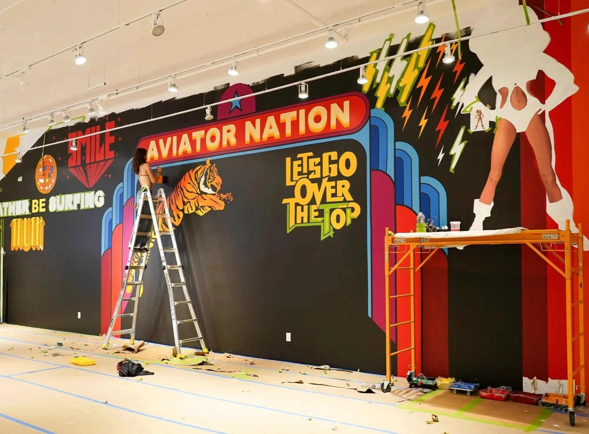 A woman standing on a ladder painting a colorful mural on a large black wall. The mural features a tiger, text that reads "Aviator Nation," "Let's Go Over the Top," and various vibrant graphics including a woman in a spacesuit, lightning bolts, and words like "Smile" and "Be Surfing."