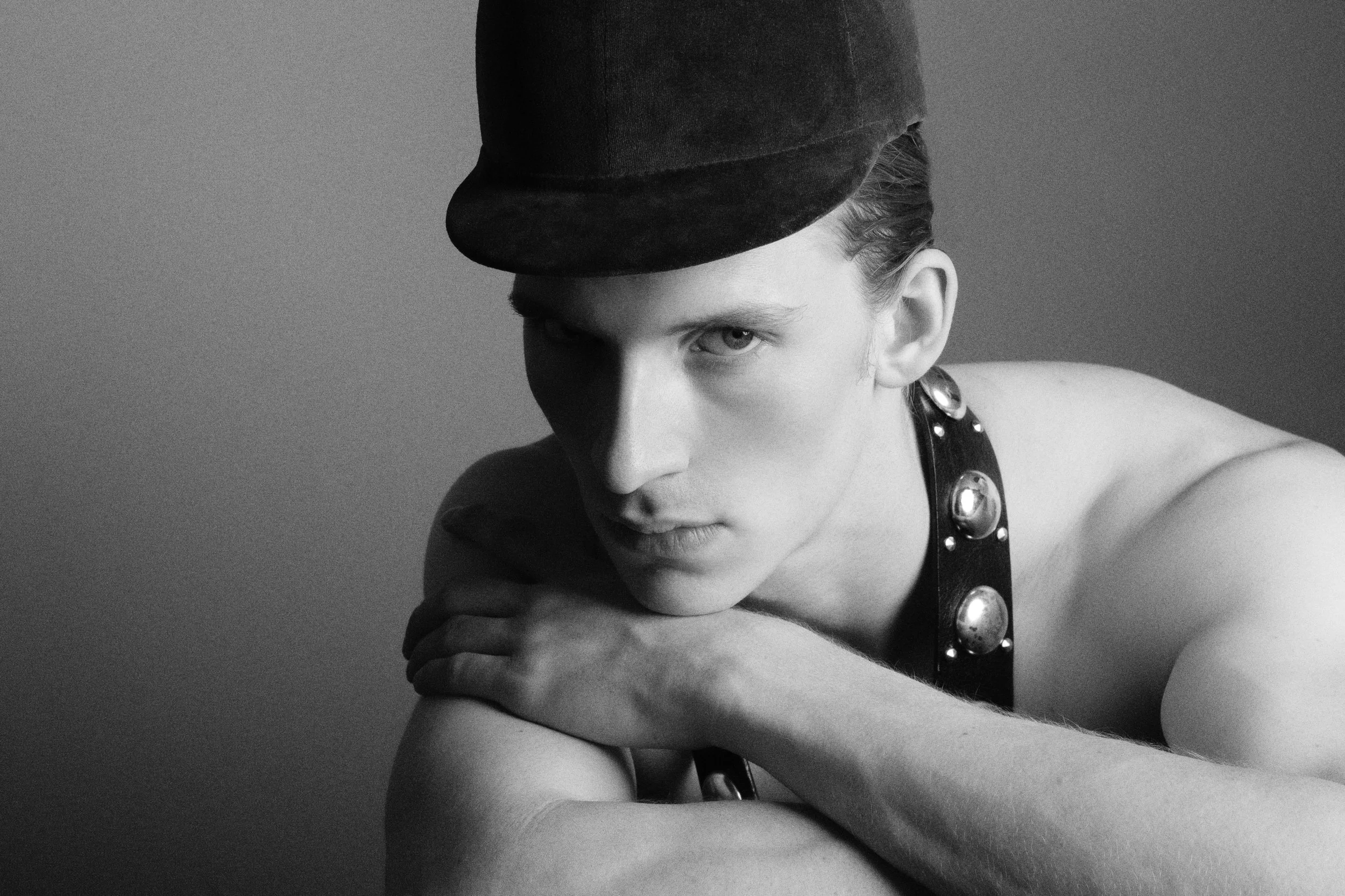 A black and white portrait of a young woman wearing a dark hat tilted forward and a sleeveless top with metallic buttons. She is resting her chin on her clasped arm, looking into the camera with a serious expression.