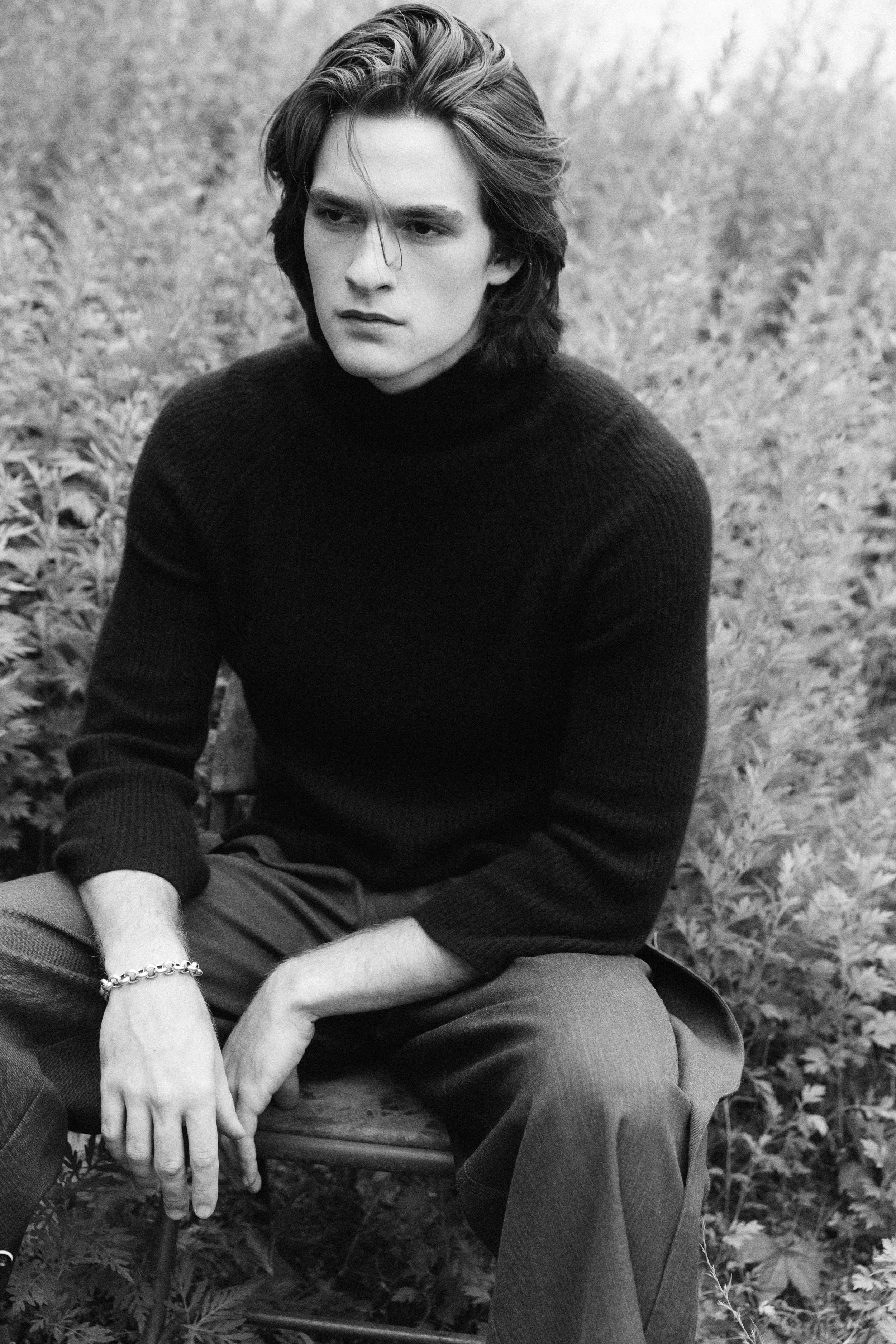 A black-and-white photo of a young man with long, wavy hair sitting outdoors among fallen leaves, wearing a dark sweater and pants, with a serious expression.