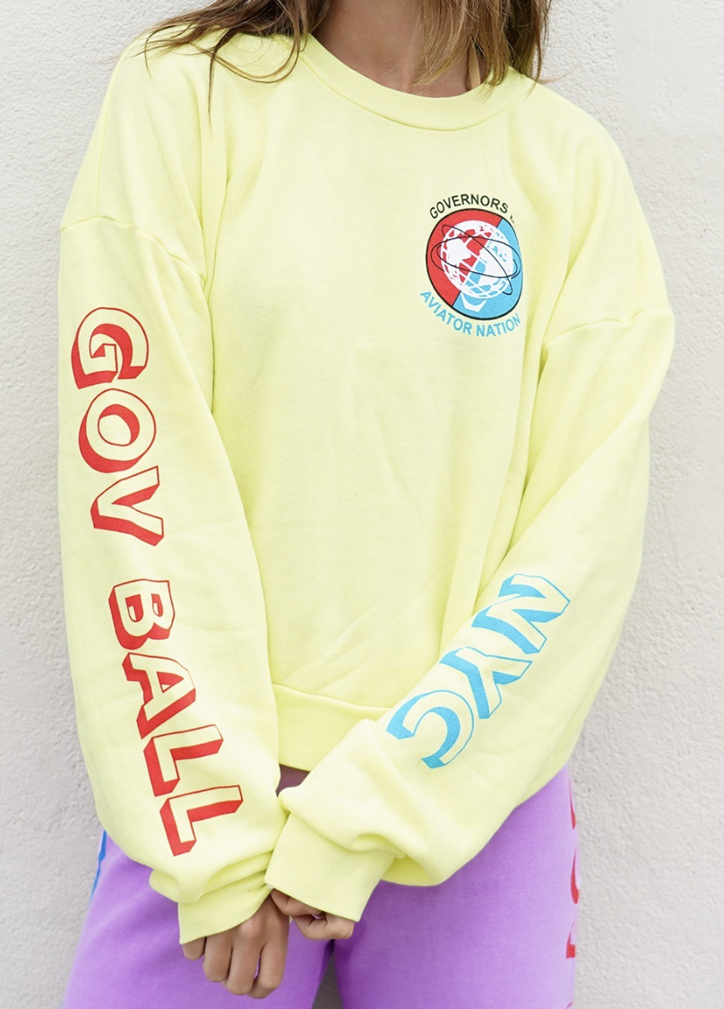 A person wearing a yellow sweatshirt with various graphic logos and text. features a graphic with the words 'GOVERNORS AVIATOR NATION' surrounding a globe design.