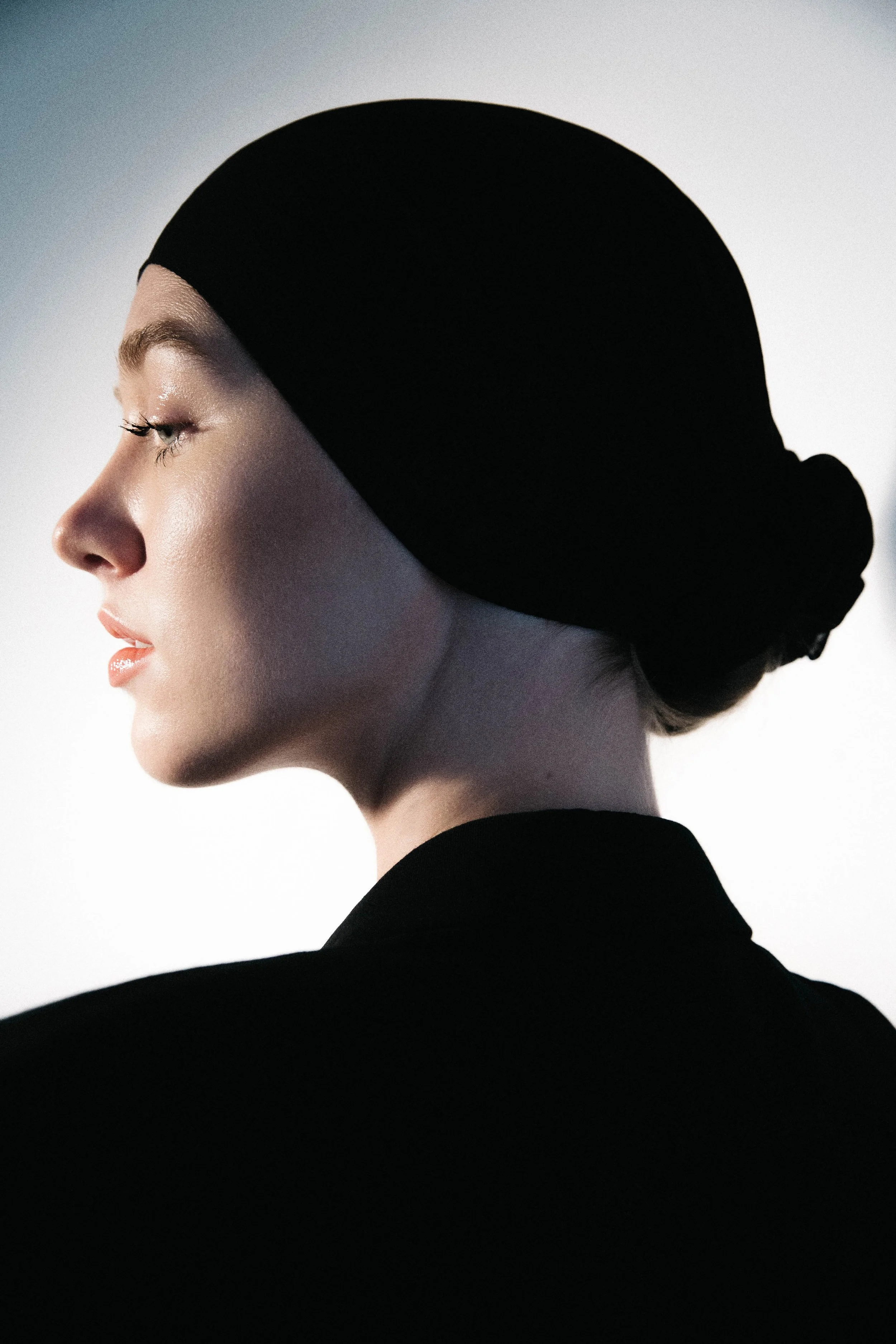 Side profile of a woman with fair skin, wearing a black headscarf and black clothing, against a gradient white and black background.