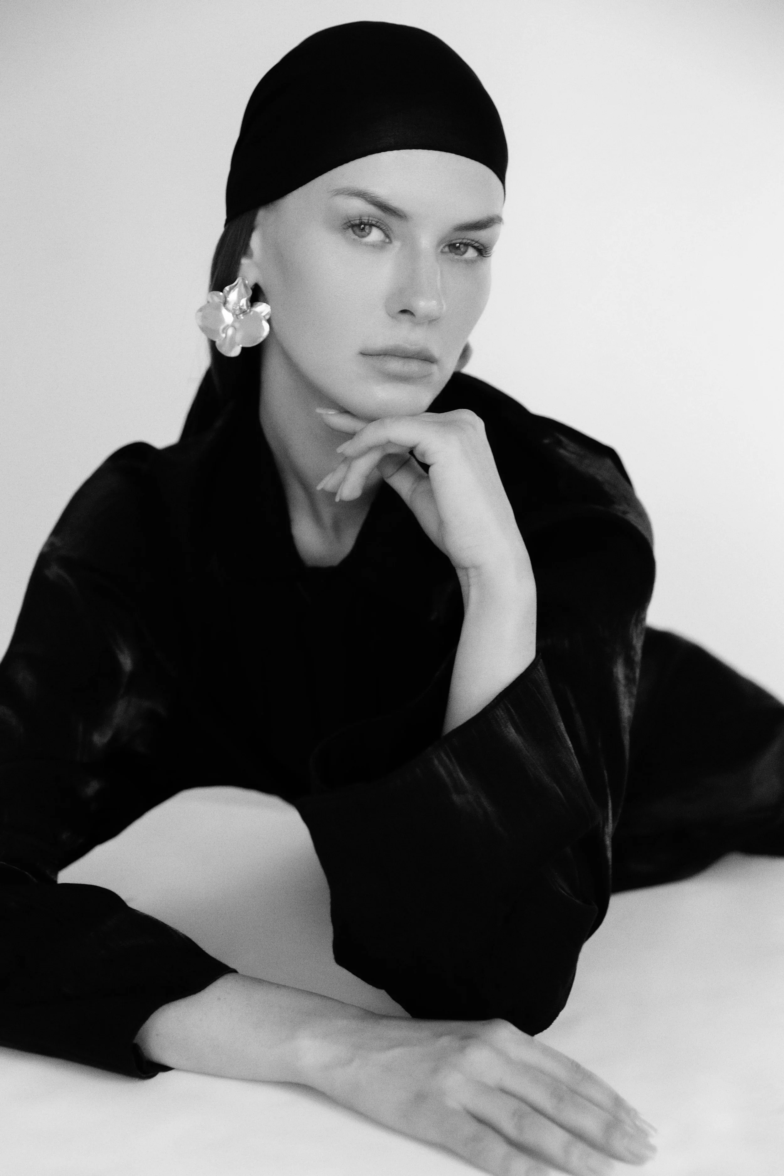 Black and white photo of a woman resting her chin on her hand, wearing a black headscarf and large flower-shaped earrings, with a focused expression.