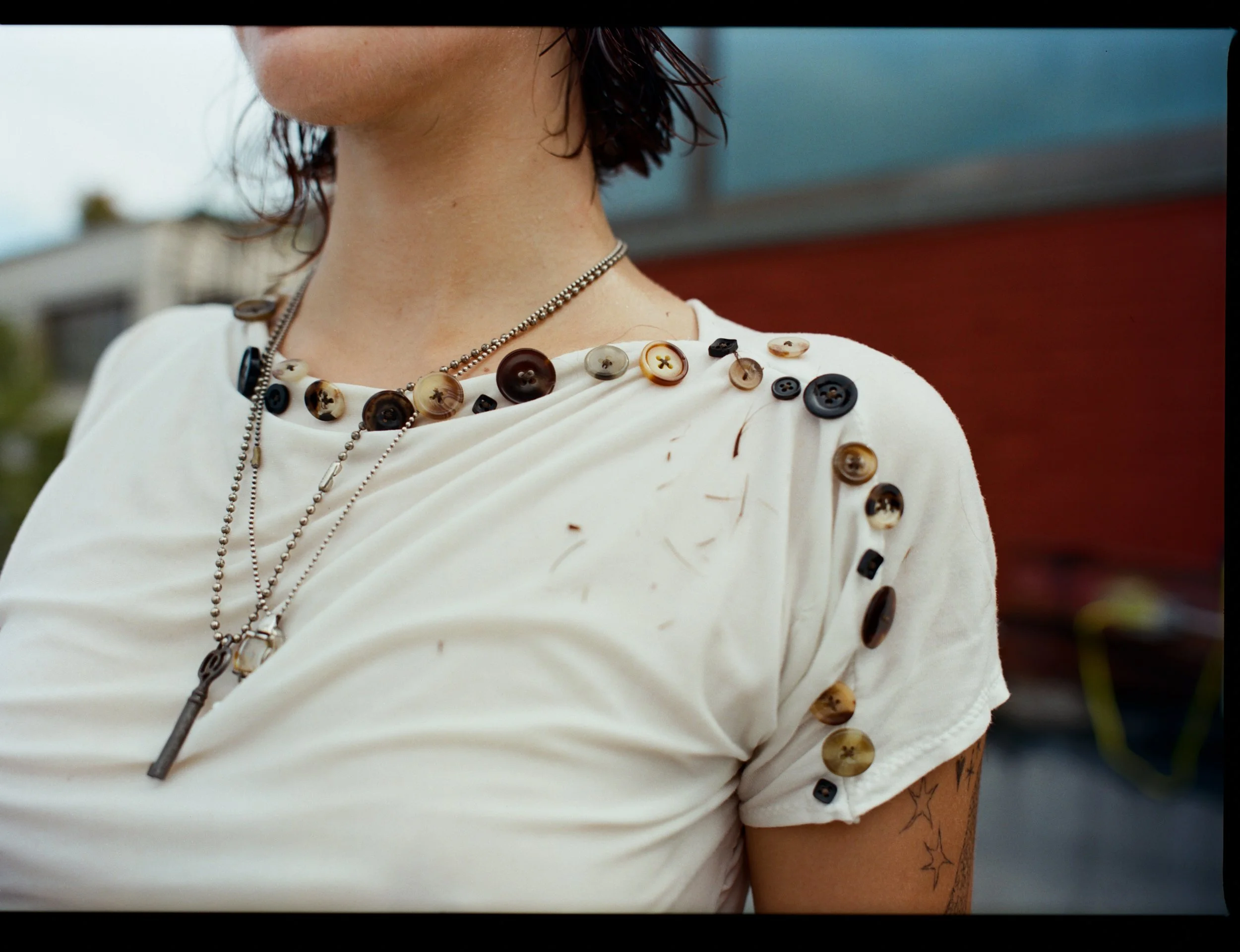 Close-up of a woman wearing a white shirt decorated with various buttons on the shoulders, a necklace with a key pendant, and a layered chain necklace. Part of her face and arm are visible, with a tattoo of stars on her upper arm.