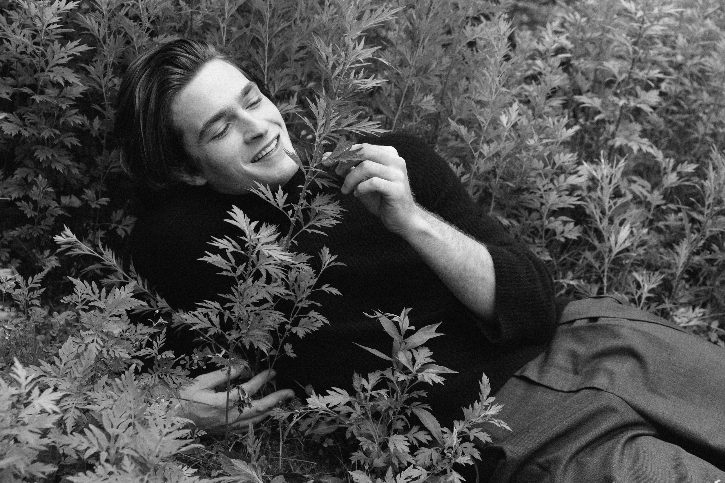 A young man lying among dense foliage, smiling with eyes closed, holding a sprig of leaves.