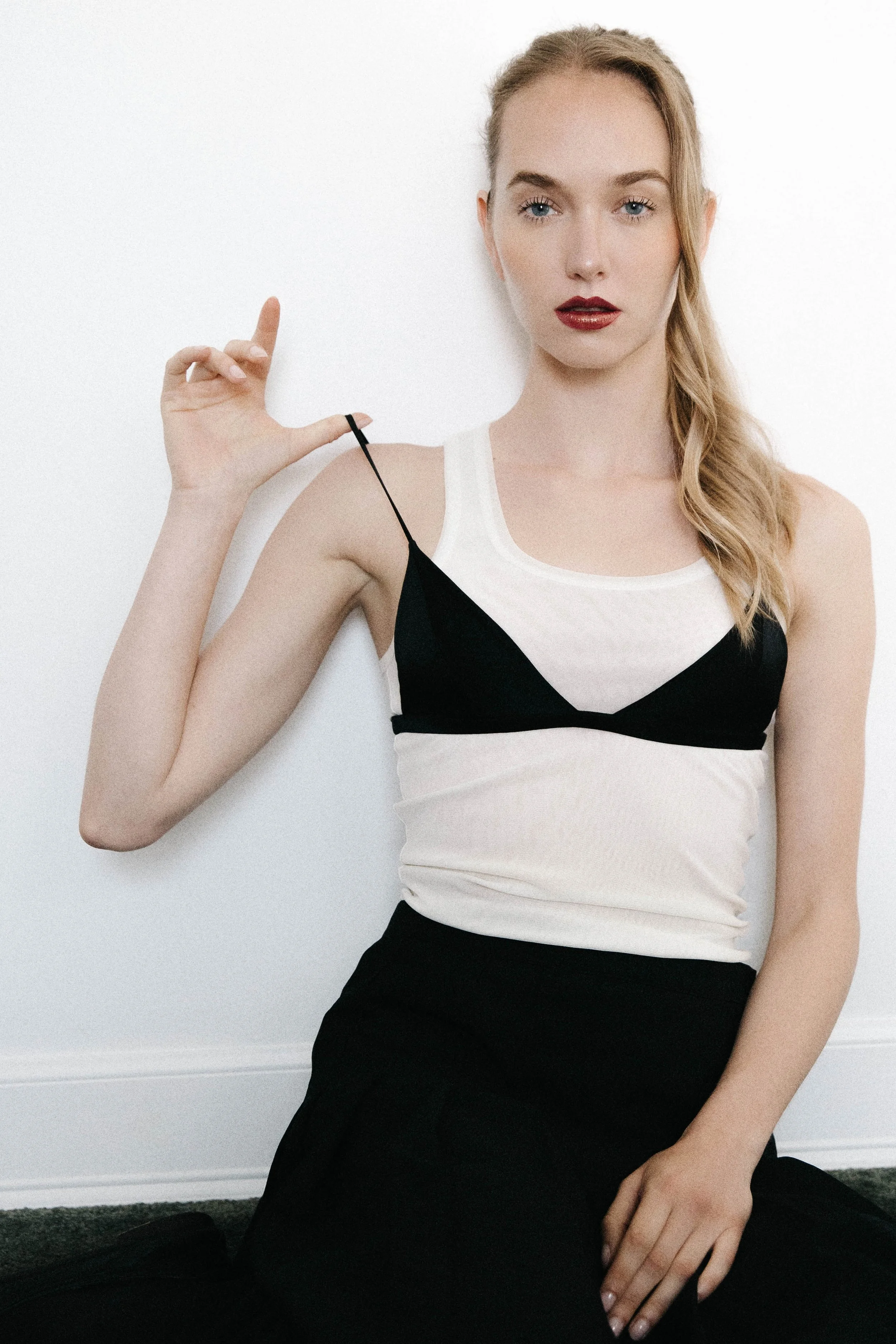 A woman with blonde hair, blue eyes, and red lipstick, sitting against a white wall, wearing a white and black sleeveless top and a black skirt, holding a black strap with her right hand.