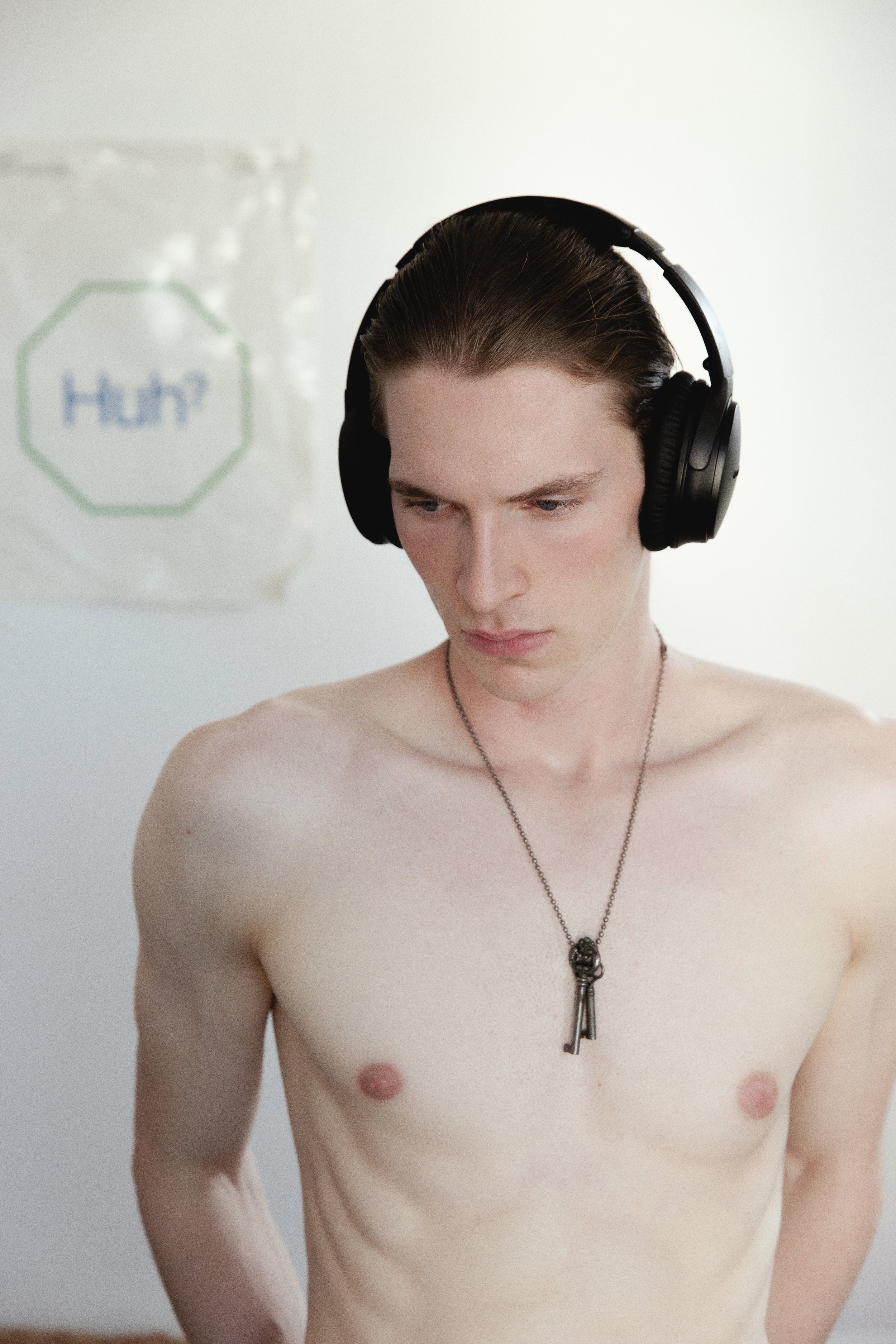 A middle-aged man with long brown hair, wearing large black over-ear headphones, is shirtless, with a necklace that has a key pendant. He is looking down with a neutral expression. The background is plain white with a bag of flour labeled 'Huh?' part