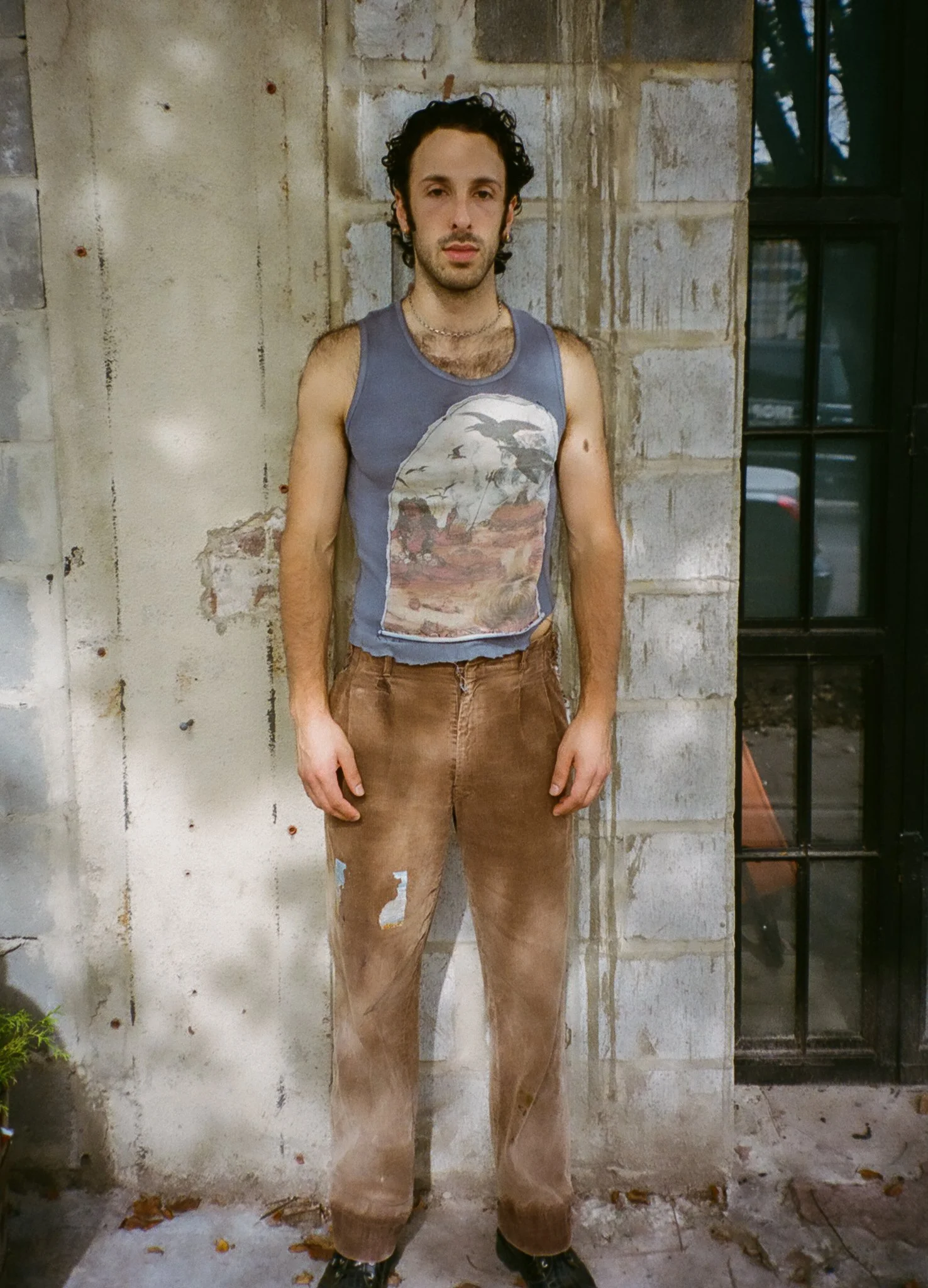 A young man with curly dark hair stands against a textured stone wall, wearing a sleeveless graphic tank top and distressed brown pants, outdoors near a window and some plants.