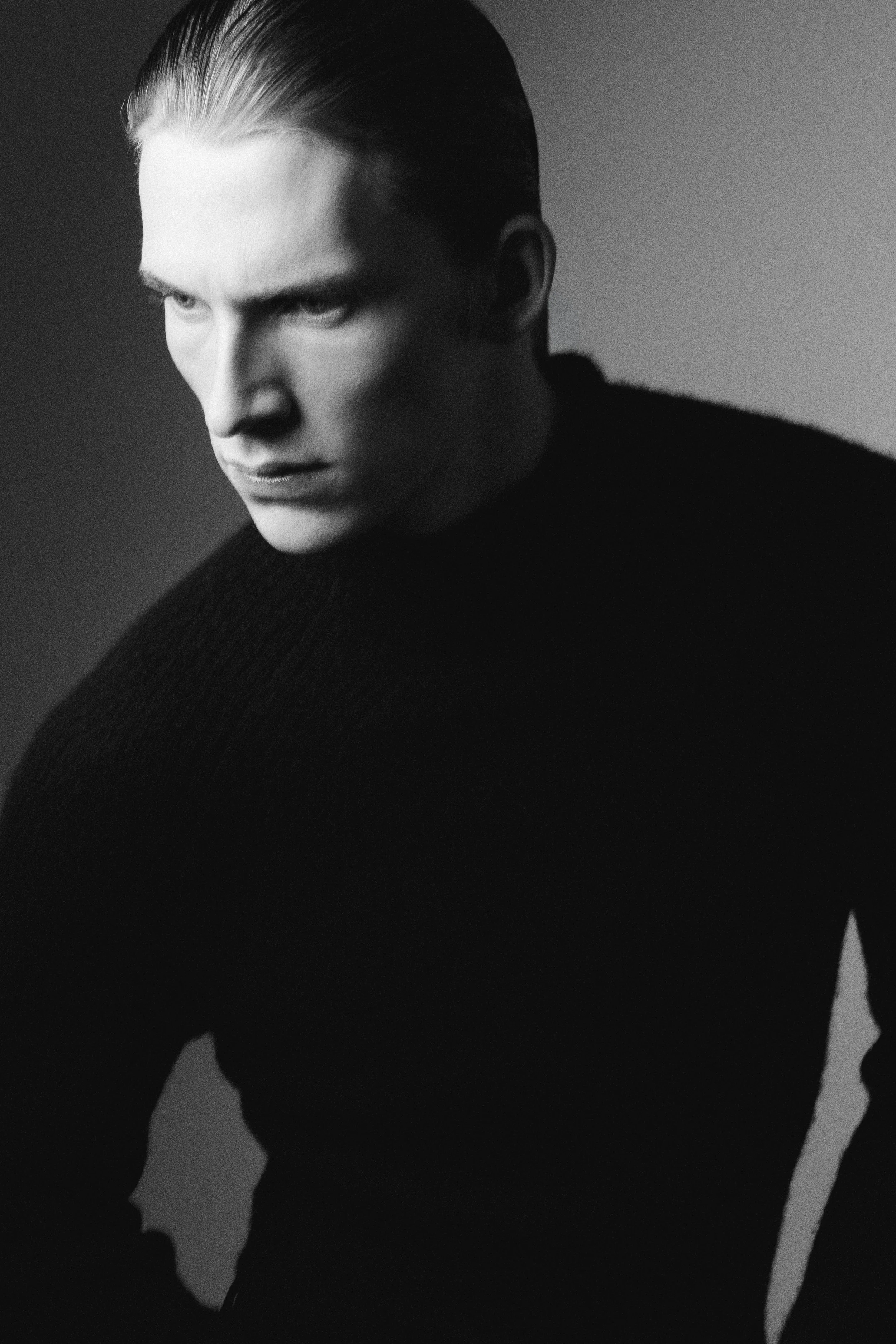 Black and white close-up image of a man with slicked-back blonde hair, wearing a dark sweater, looking down and away from the camera, with a serious expression.