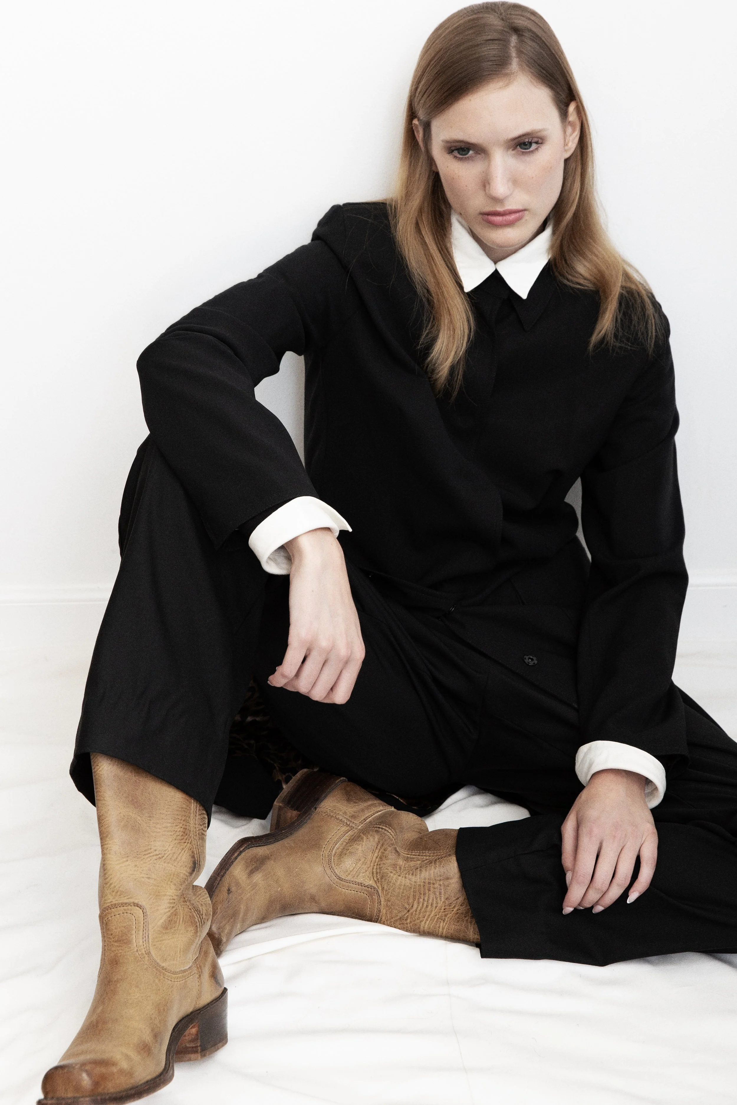 A red-haired woman wearing a black sweater with white collar cuffs and black pants, sitting with her legs crossed, wearing tan cowboy boots, against a plain white background.