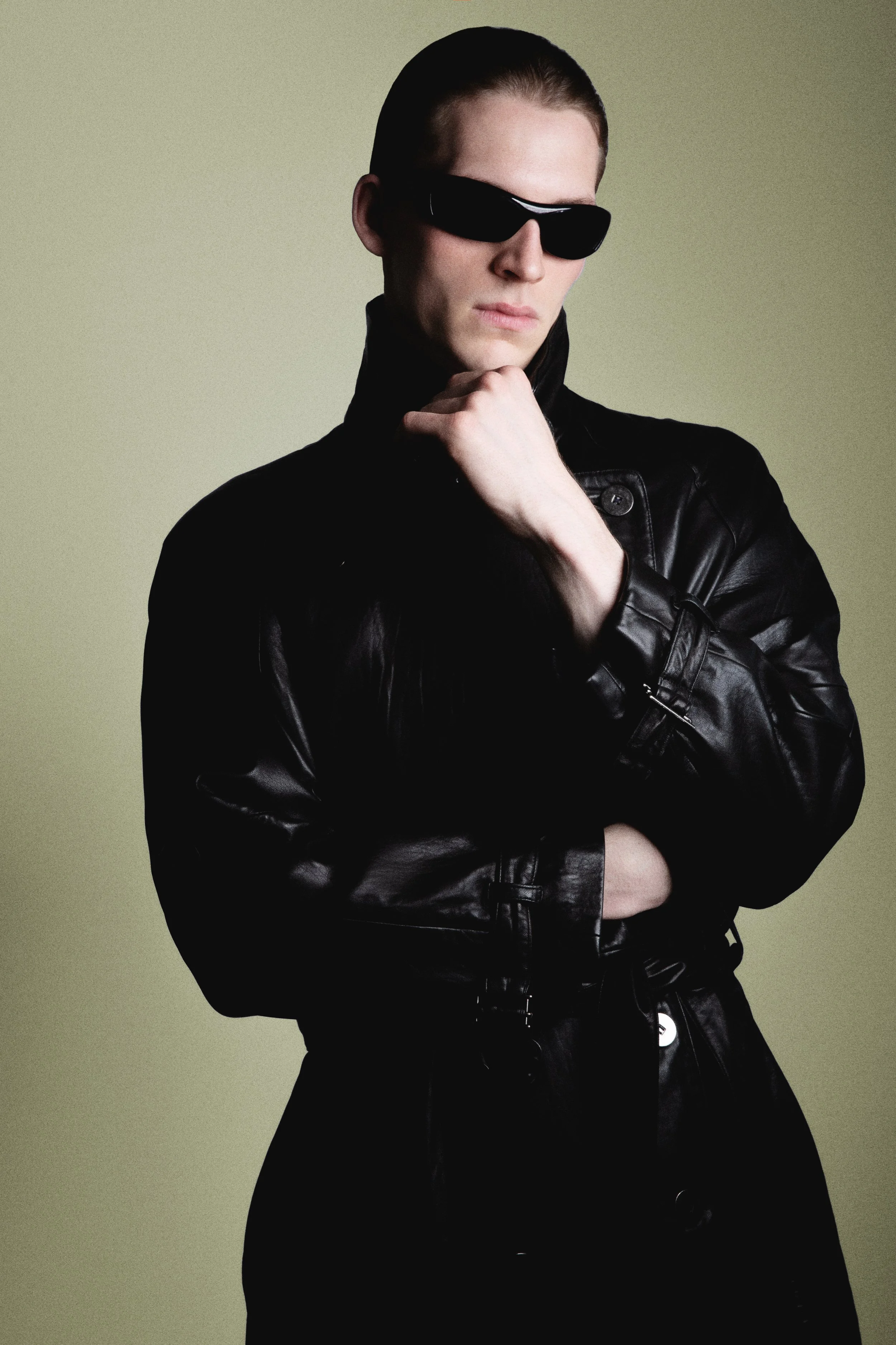 A person wearing a black leather jacket and black sunglasses, with hair pulled back, standing with one hand on their chin and the other crossed over their chest against a plain, light-colored background.