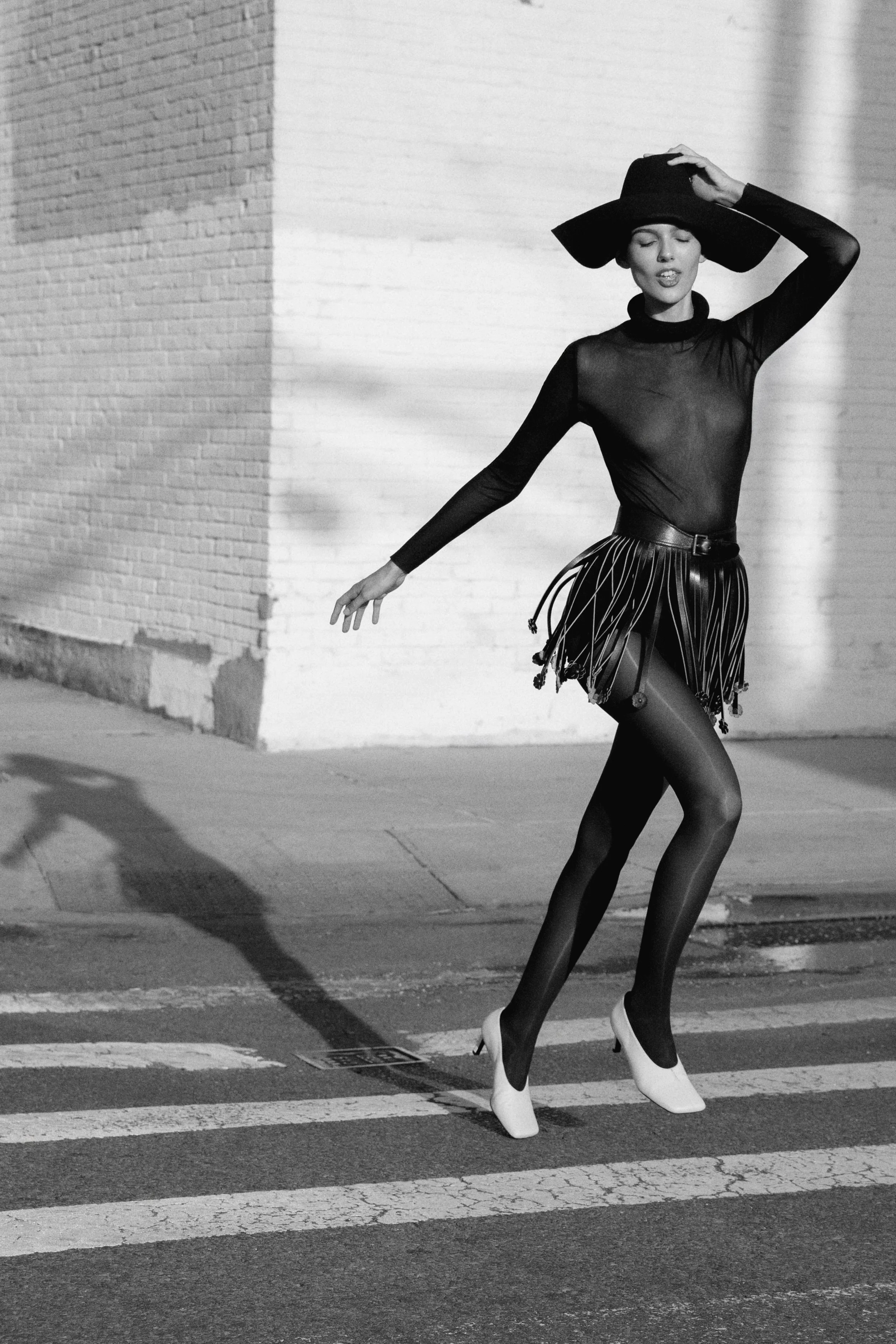 A woman in high-heeled boots and a sheer top, wearing a wide-brimmed hat, posing on a street in front of a brick wall, with one hand on her head.