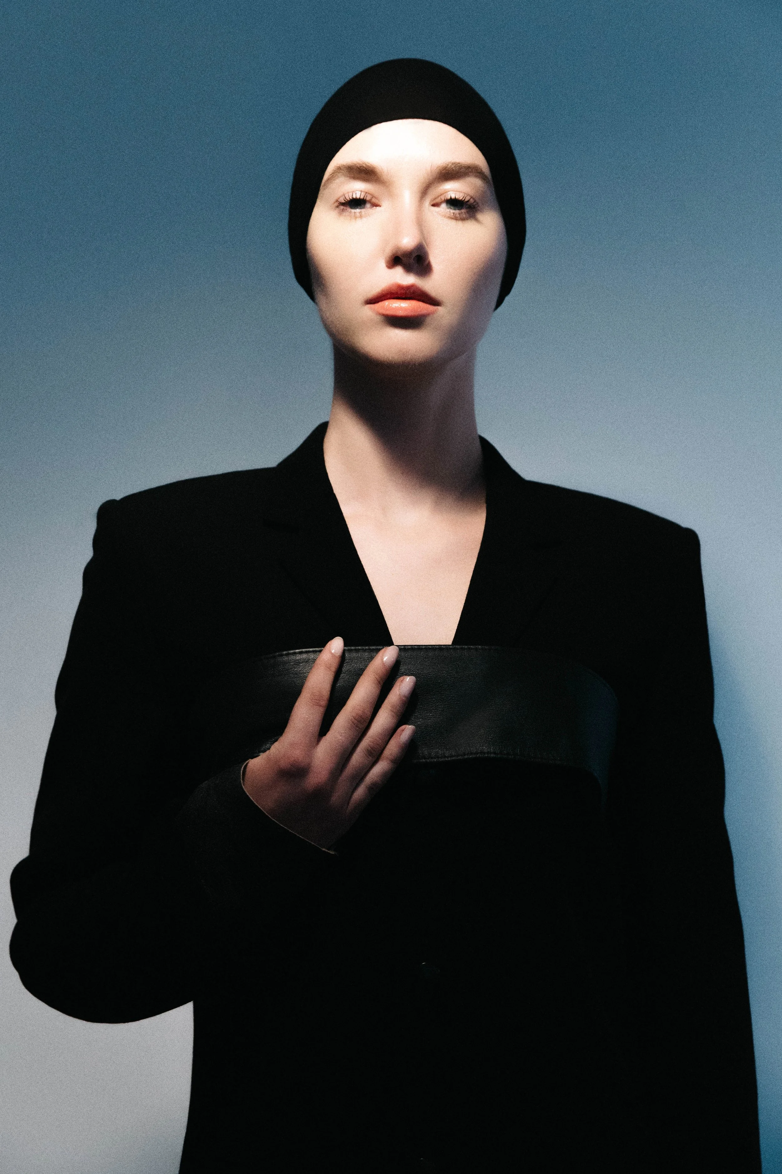 A woman with a fair complexion and red lips wearing a black beanie and black outfit, holding a black leather clutch, standing against a gradient background transitioning from blue to gray.