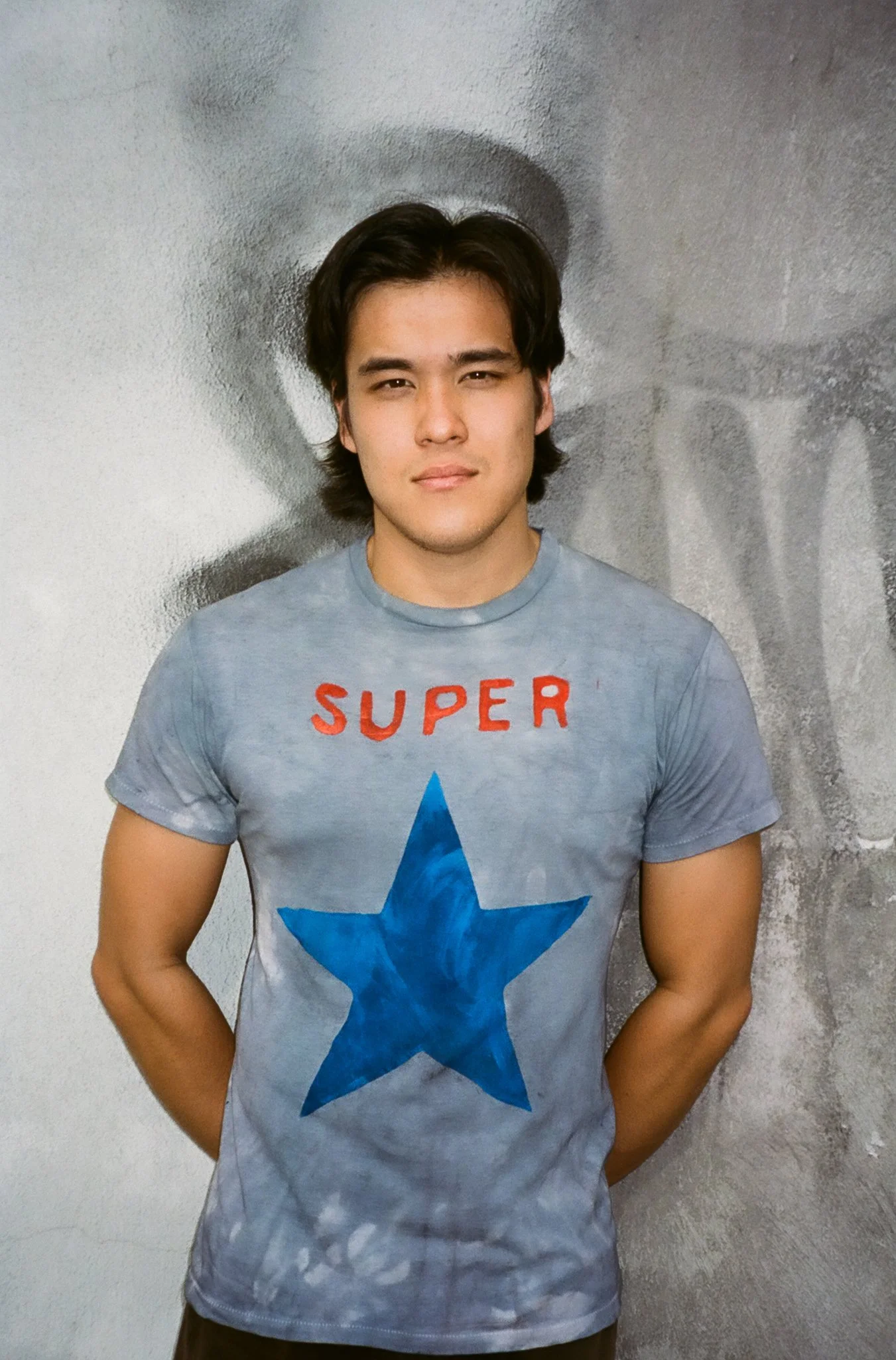 Young man with dark hair wearing a light gray T-shirt with a blue star and the word 'SUPER' in red on the front, standing against a textured wall.