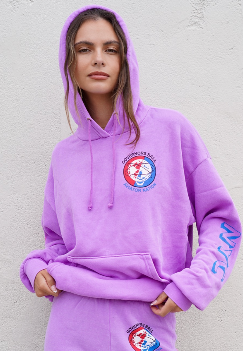 A woman wearing a purple hoodie and matching sweatpants, with logos for Governers Ball and Aviator Nation on the hoodie, standing against a plain wall.