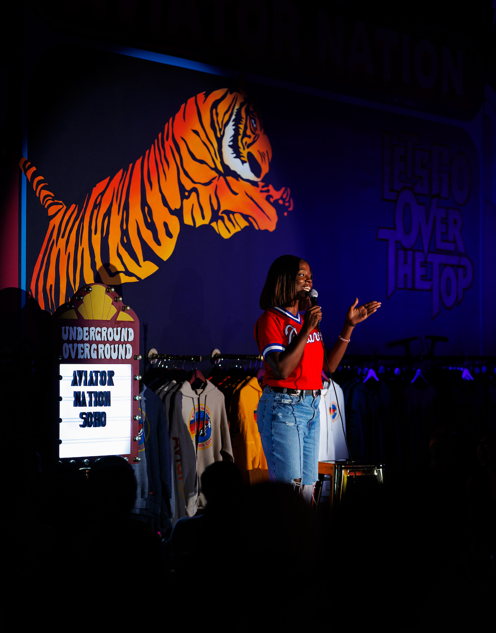 A woman speaking into a microphone on stage at a retail store, with a large orange tiger graphic on the wall behind her and clothing racks with colorful hoodies. The store sign reads 'Underground Overground' and a sign with the words 'Let Go Over The Top' is visible in the background.