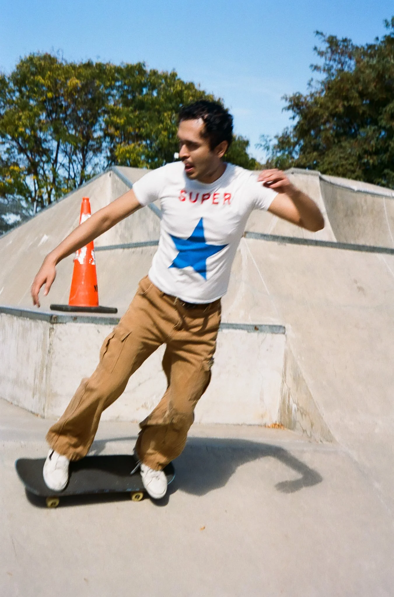 A man skateboarding at a skate park on a sunny day, wearing a white T-shirt with a blue star and red text, tan cargo pants, and white shoes.