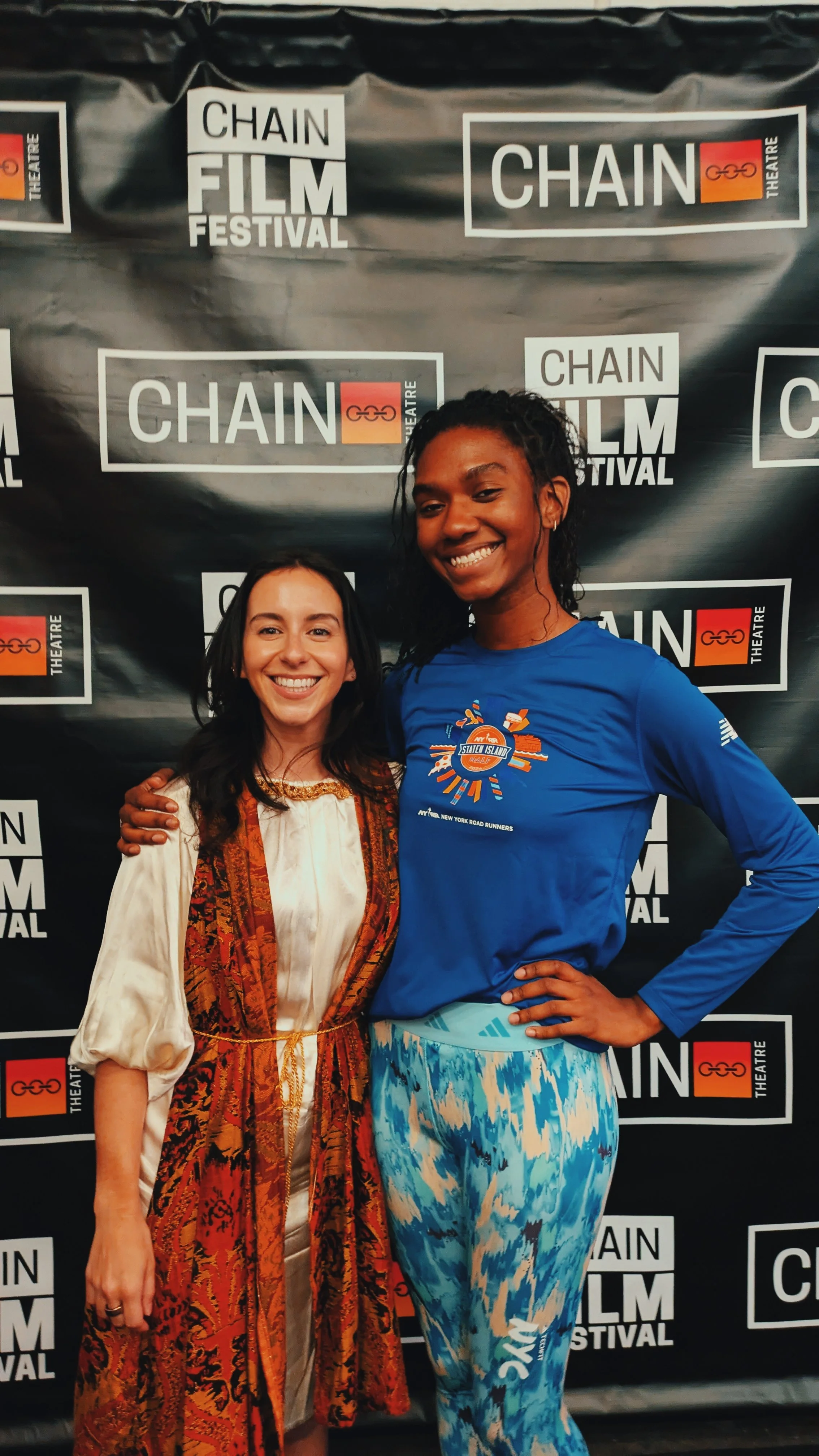 Two women smiling at a film festival, standing in front of a step and repeat banner with 'CHAIN FILM FESTIVAL' and 'CHAIN THEATRE' logos.