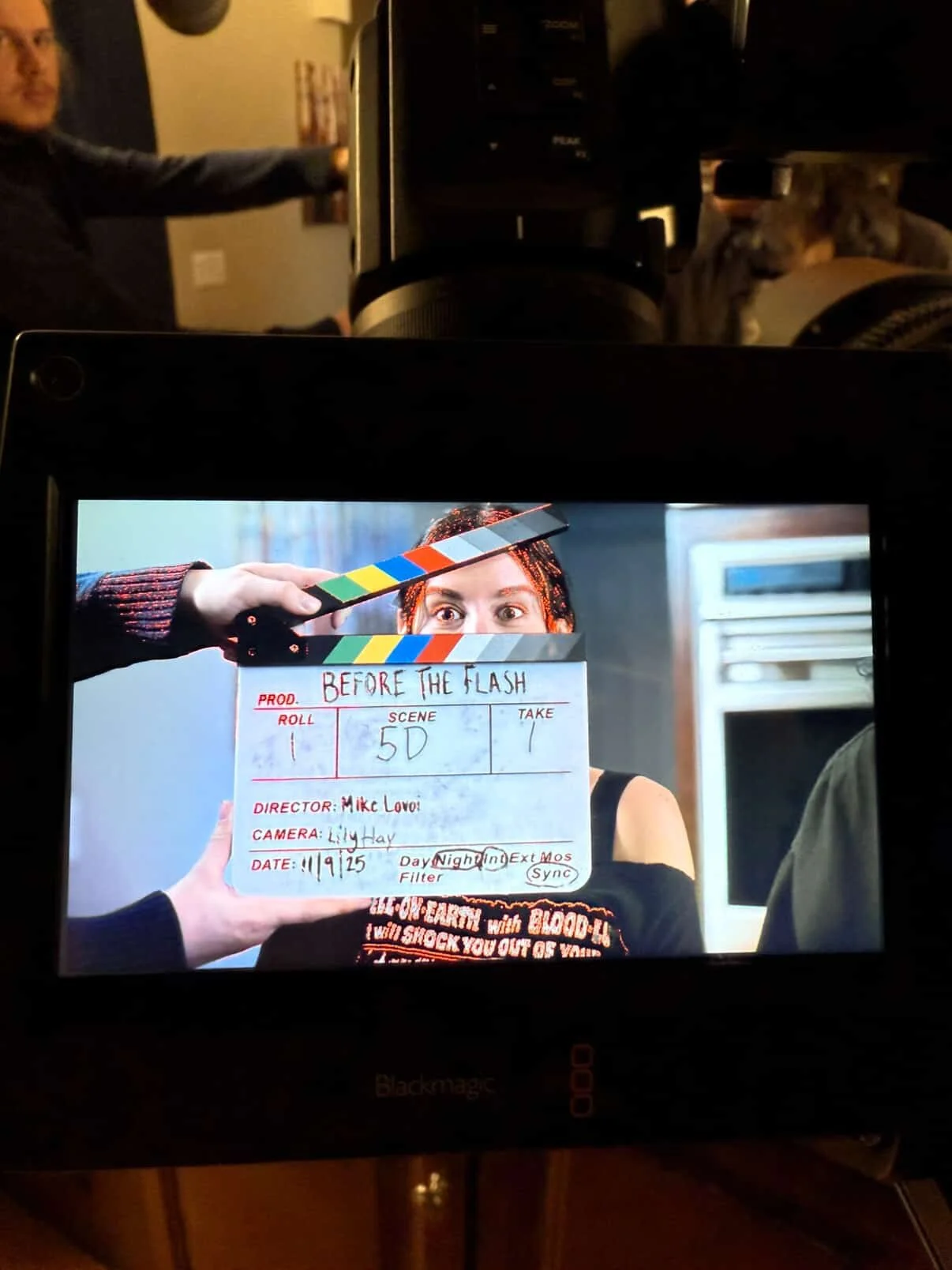 A television screen shows a film production scene with a clapperboard, a woman with red hair, and a person in the background with a dog. The clapperboard reads "BEFORE THE FLASH," dated 11/9/15, with various production details.
