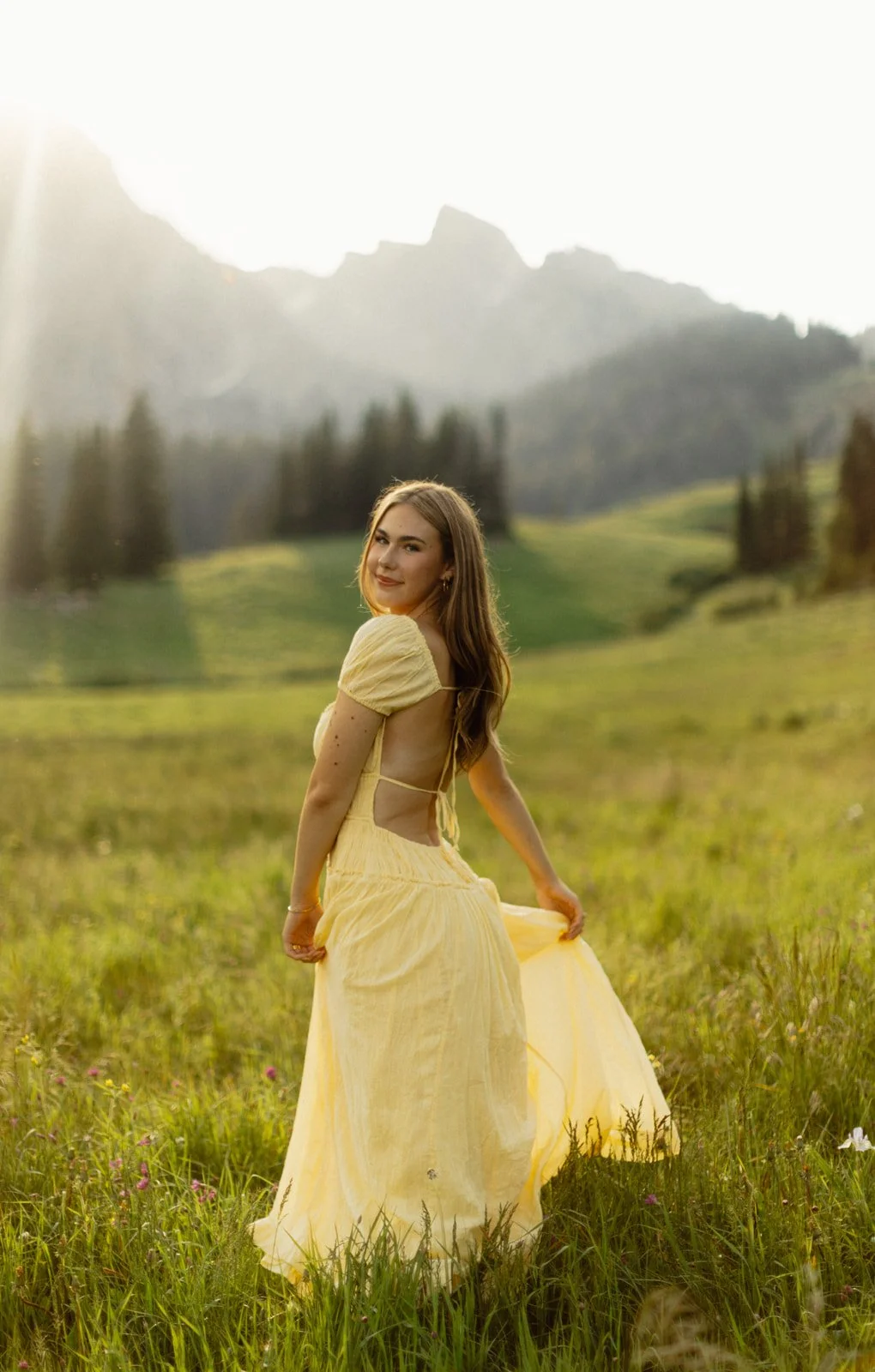 Young woman in a yellow dress standing in a grassy field with mountains and trees in the background, holding the edge of her dress and smiling.
