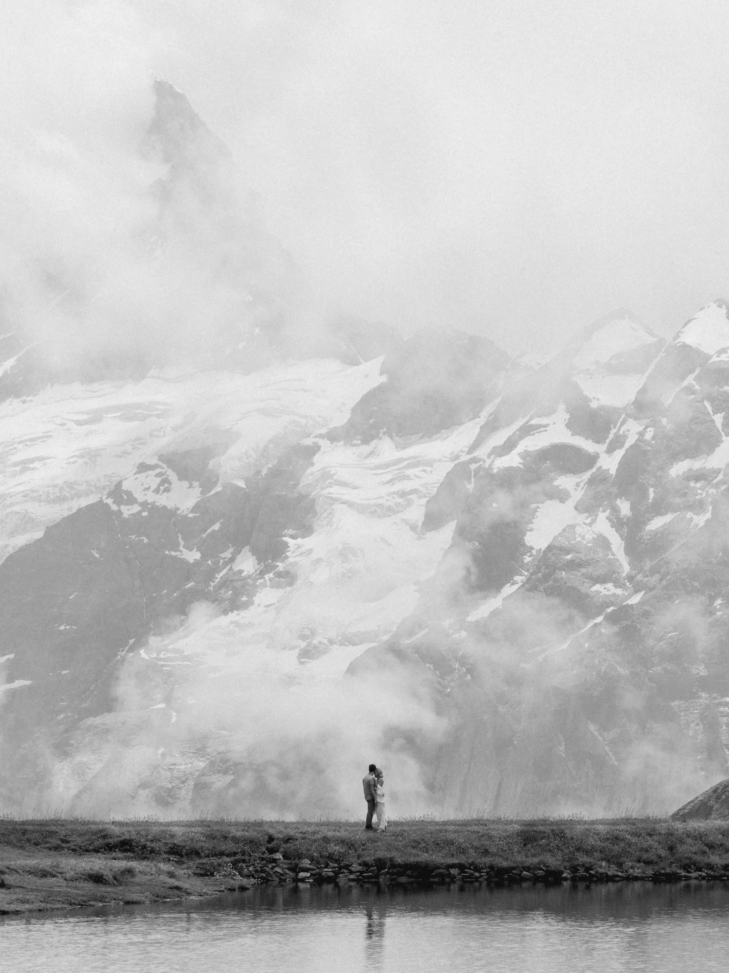 A black-and-white photo of a couple standing by a body of water with snow-capped mountains and mist in the background.