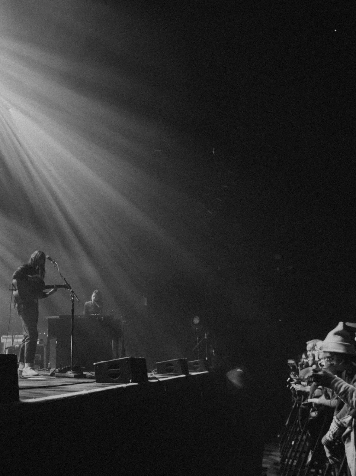 Black and white photo of a live music concert with two musicians on stage, one playing guitar and the other at a keyboard. Audience members are visible on the right side, some taking photos and videos.