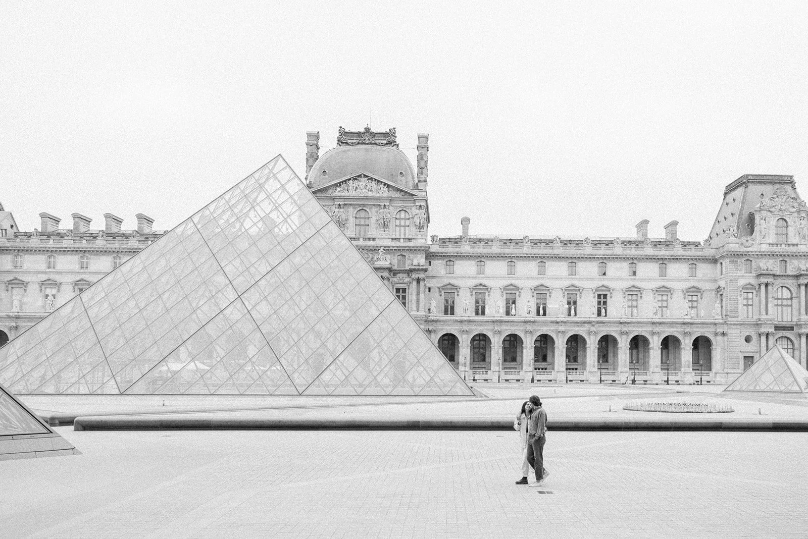 A black-and-white photo of the Louvre Museum in Paris with modern glass pyramids in the foreground and a historic building in the background. Two people stand close together, talking in the open courtyard.