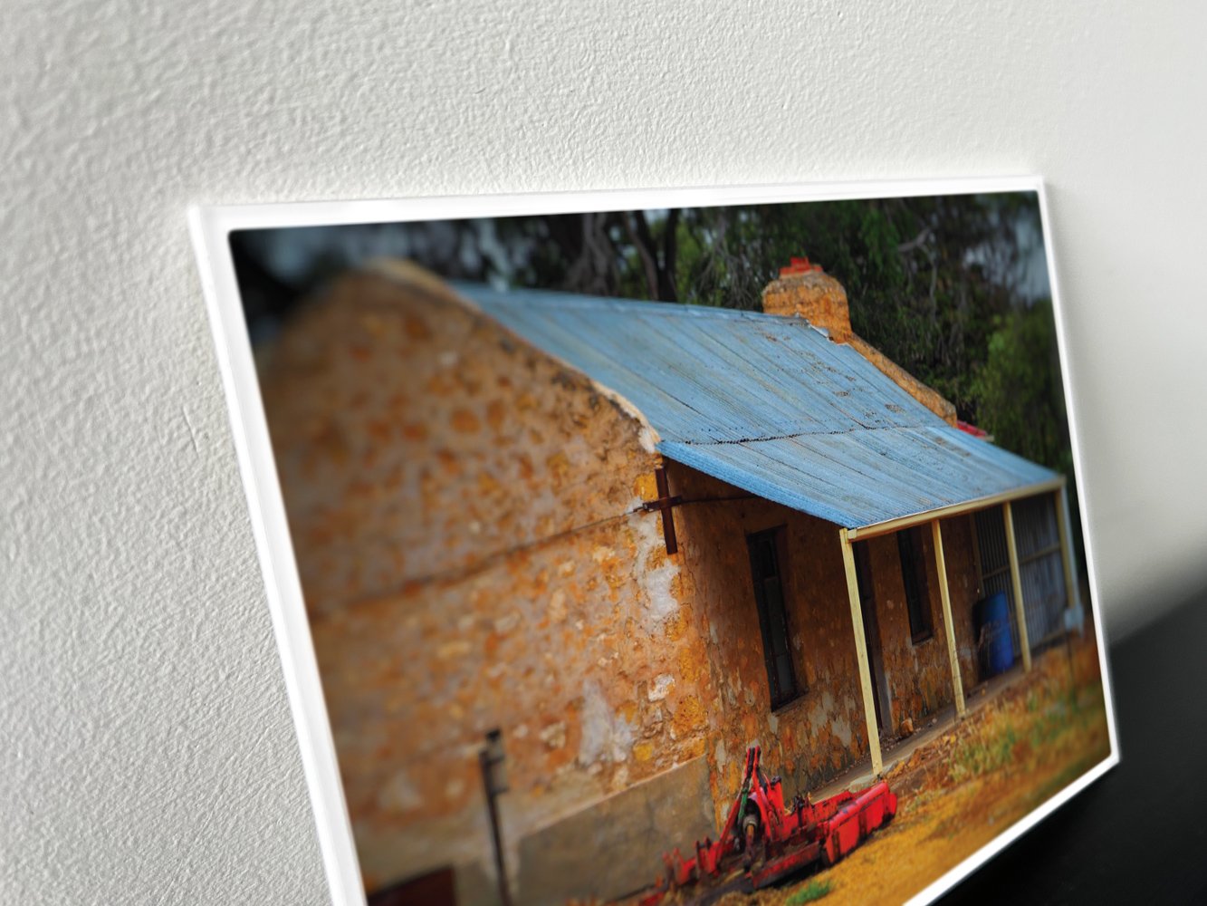 Metal backed photograph of an old stone house with a corrugated metal roof and a wooden porch in country Australia. A red farming implement is in the front yard.