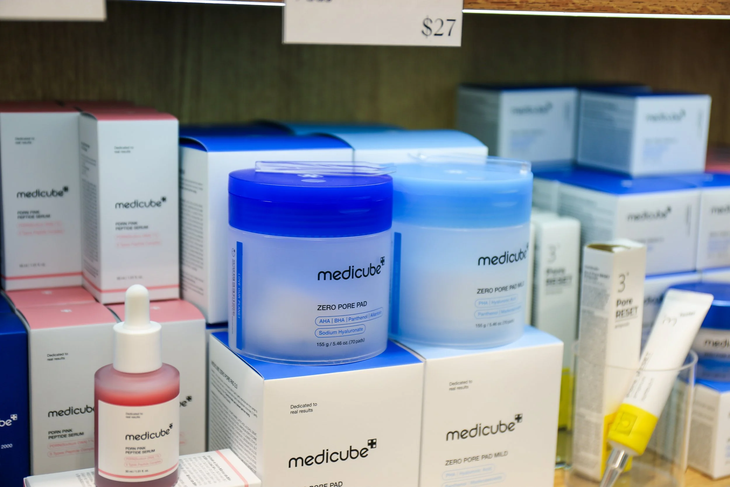 Display of skincare products including blue containers labeled 'medicube' with Zero Pore Pad, skincare serum in a pink bottle, and various other skincare boxes on a shelf.
