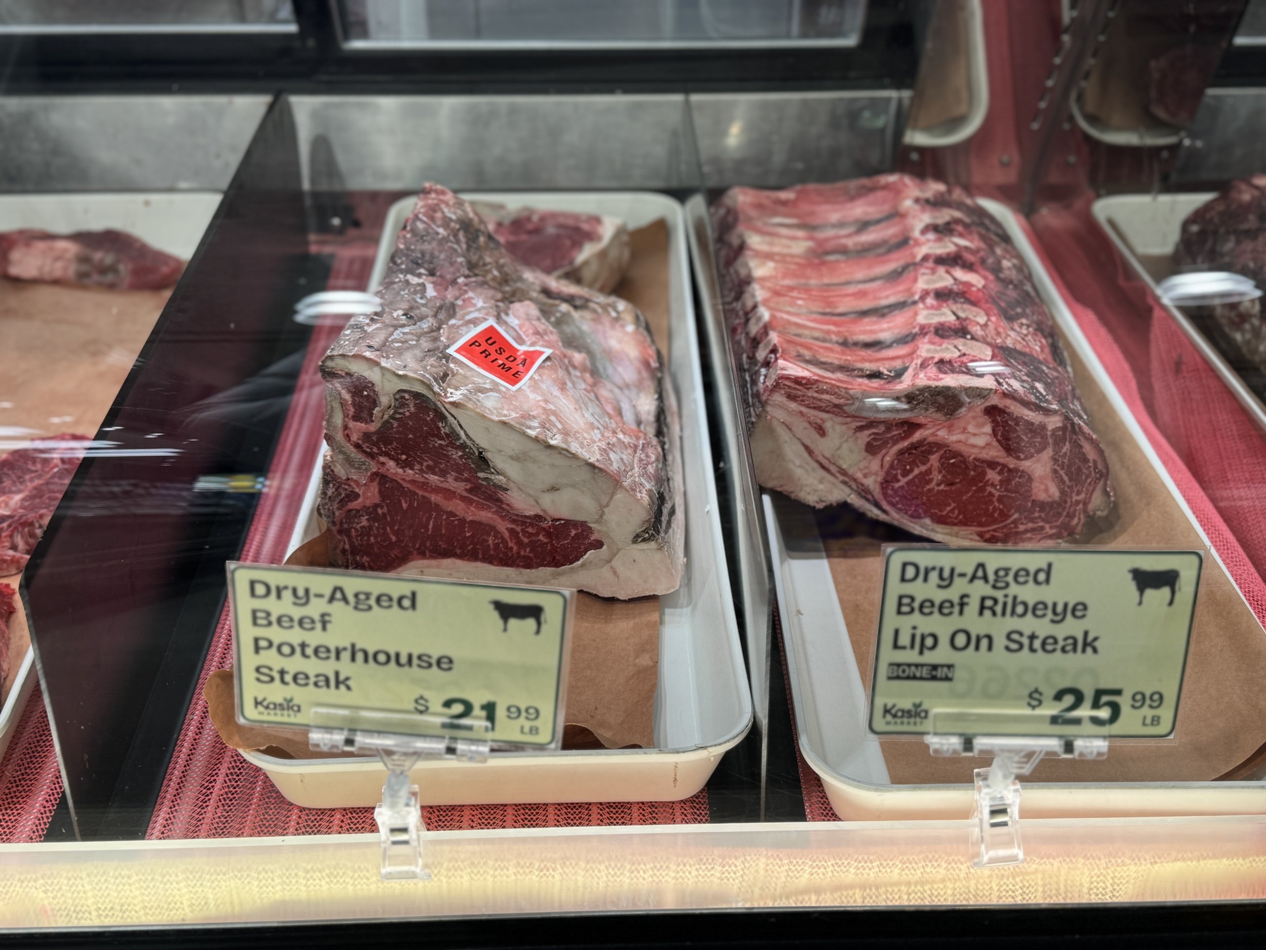 Showcase of dry-aged beef cuts, including Poterhouse Steak on the left and Ribeye Lip On Steak on the right, in a refrigerated display case at a butcher shop. The Poterhouse Steak is labeled at $21.99 per pound, and the Ribeye Lip On Steak is priced at $25.99 per pound.