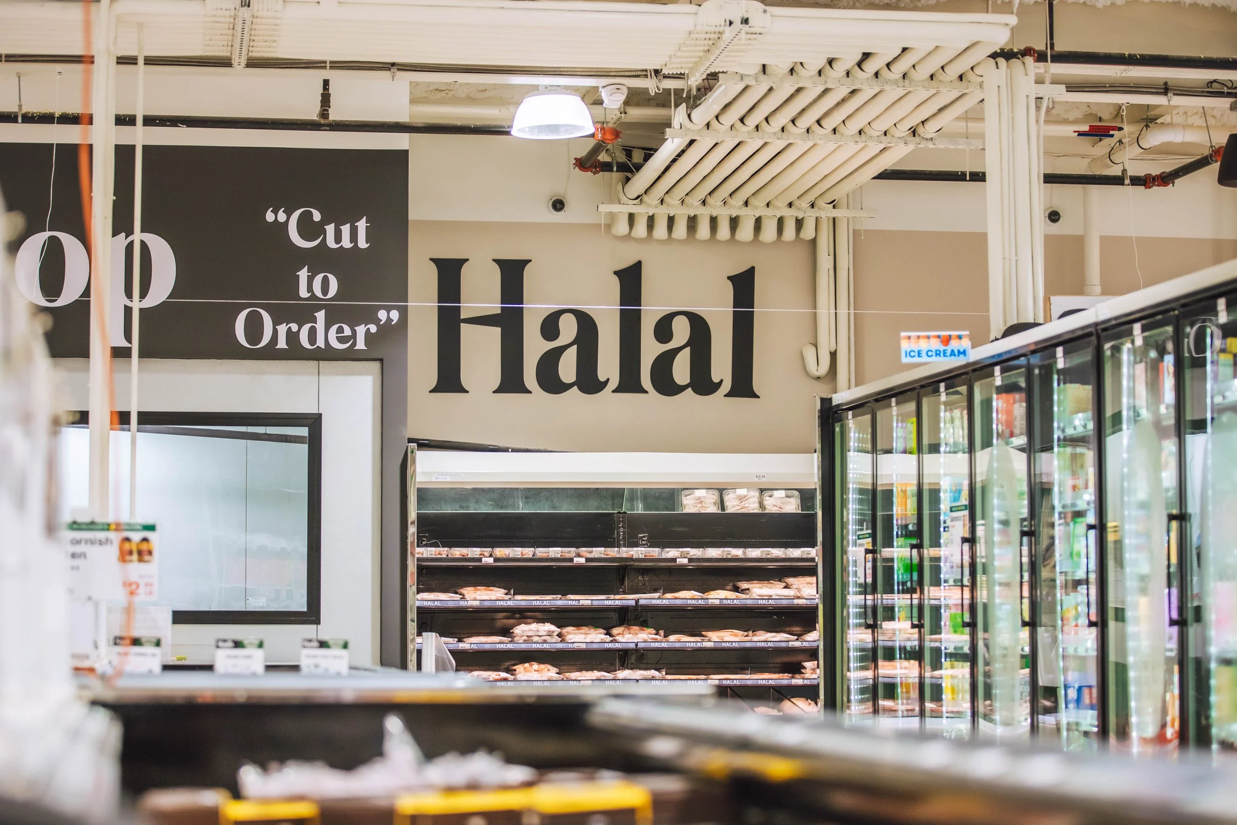 Grocery store refrigerated section with a large sign reading 'Halal' and 'Cut to Order', with frozen meat and ice cream visible.