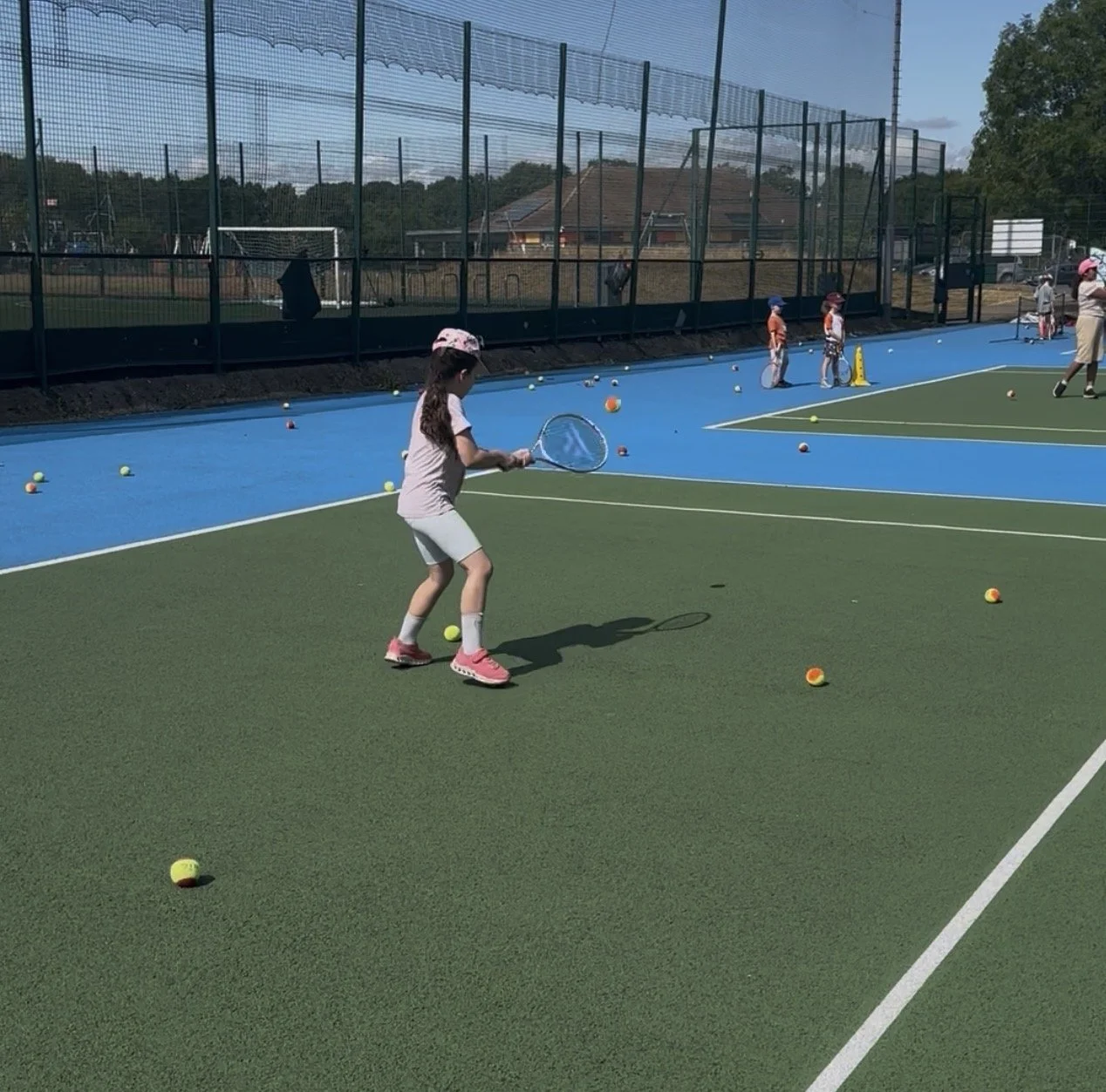 Children practicing tennis on a multi-sport court, with tennis balls scattered across the court, some kids holding tennis rackets, and others standing near cones.