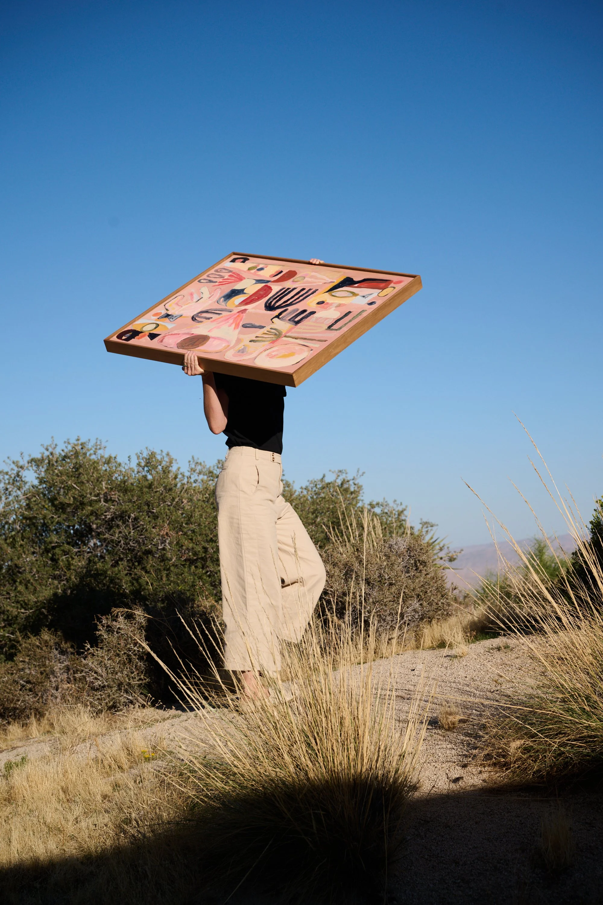 Person standing outdoors in a desert-like area, holding a large, colorful abstract painting above their head. The person is wearing beige pants and a black shirt, with a clear blue sky and shrubbery in the background.