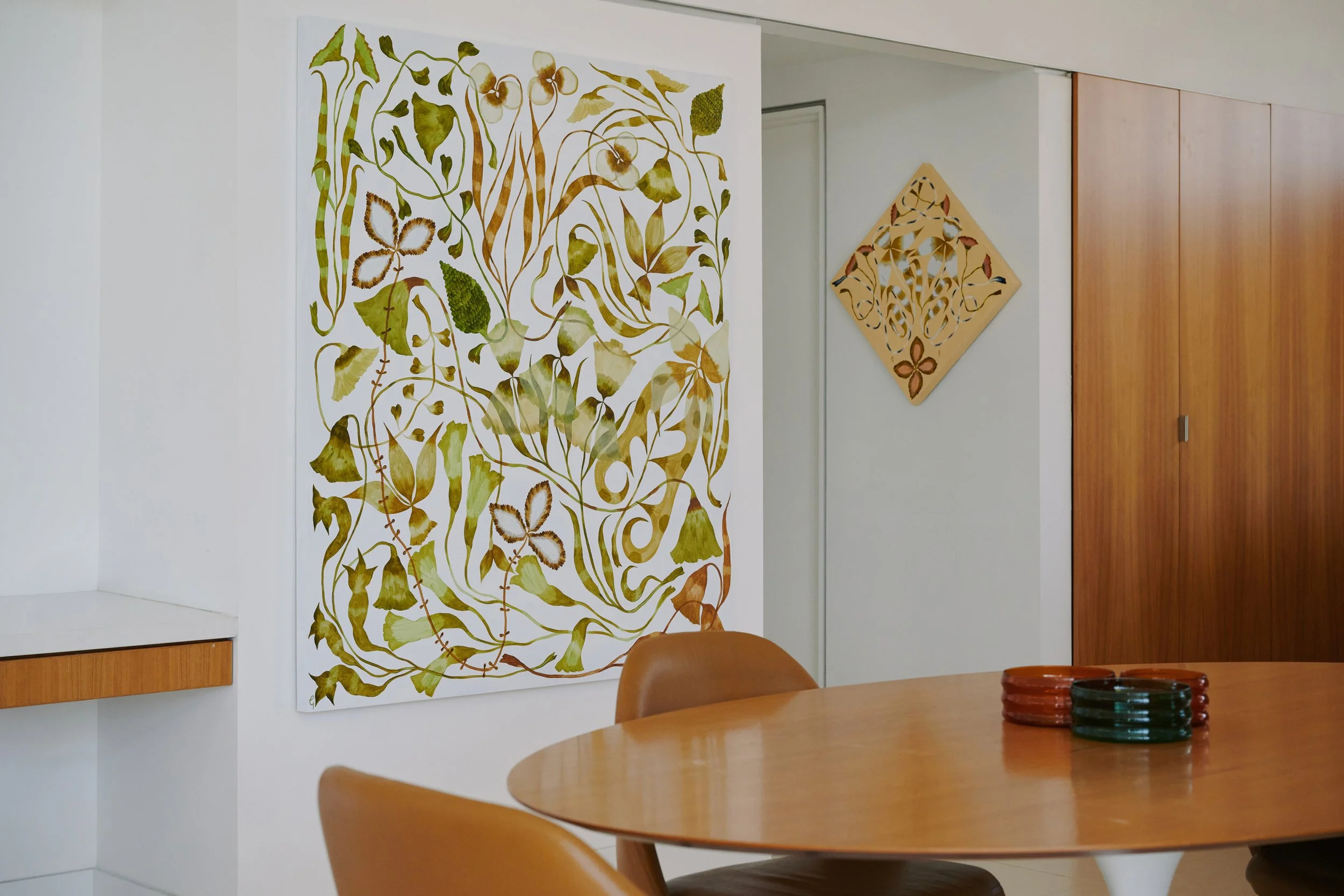 A modern dining area with a round wooden table, brown chairs, and decorative artwork on the walls featuring botanical themes.