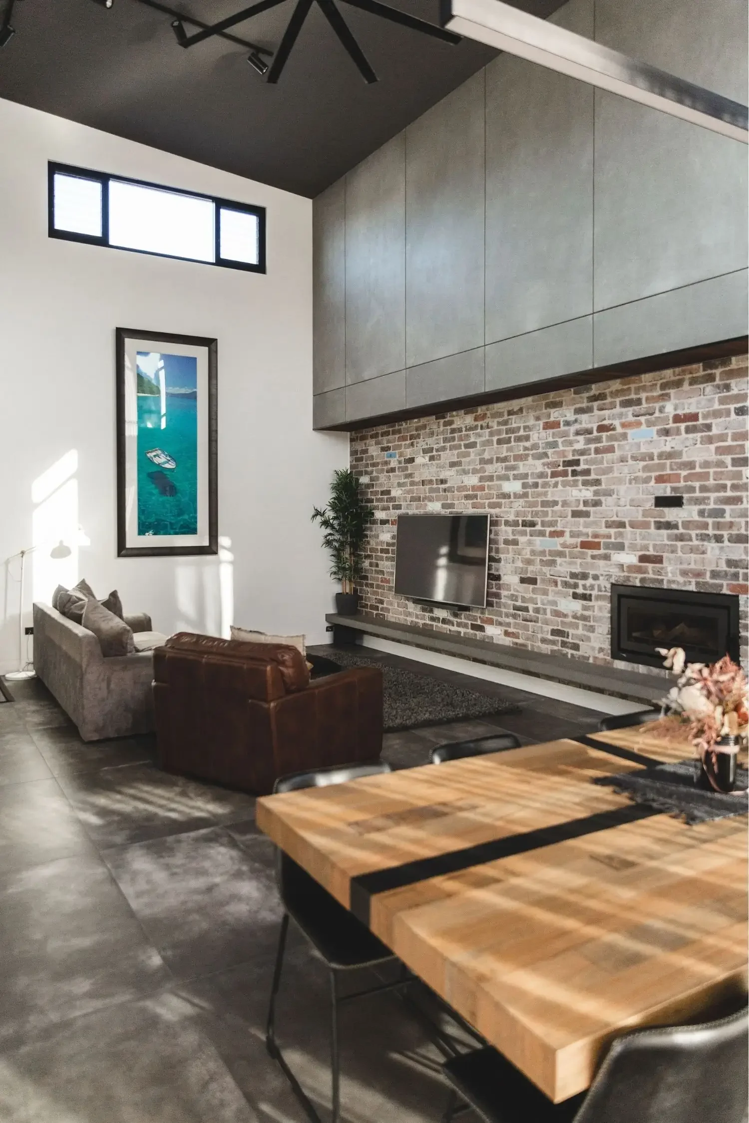 Modern living room with high vaulted ceiling, brick accent wall with TV, gray and brown furniture, large window, and contemporary decor.