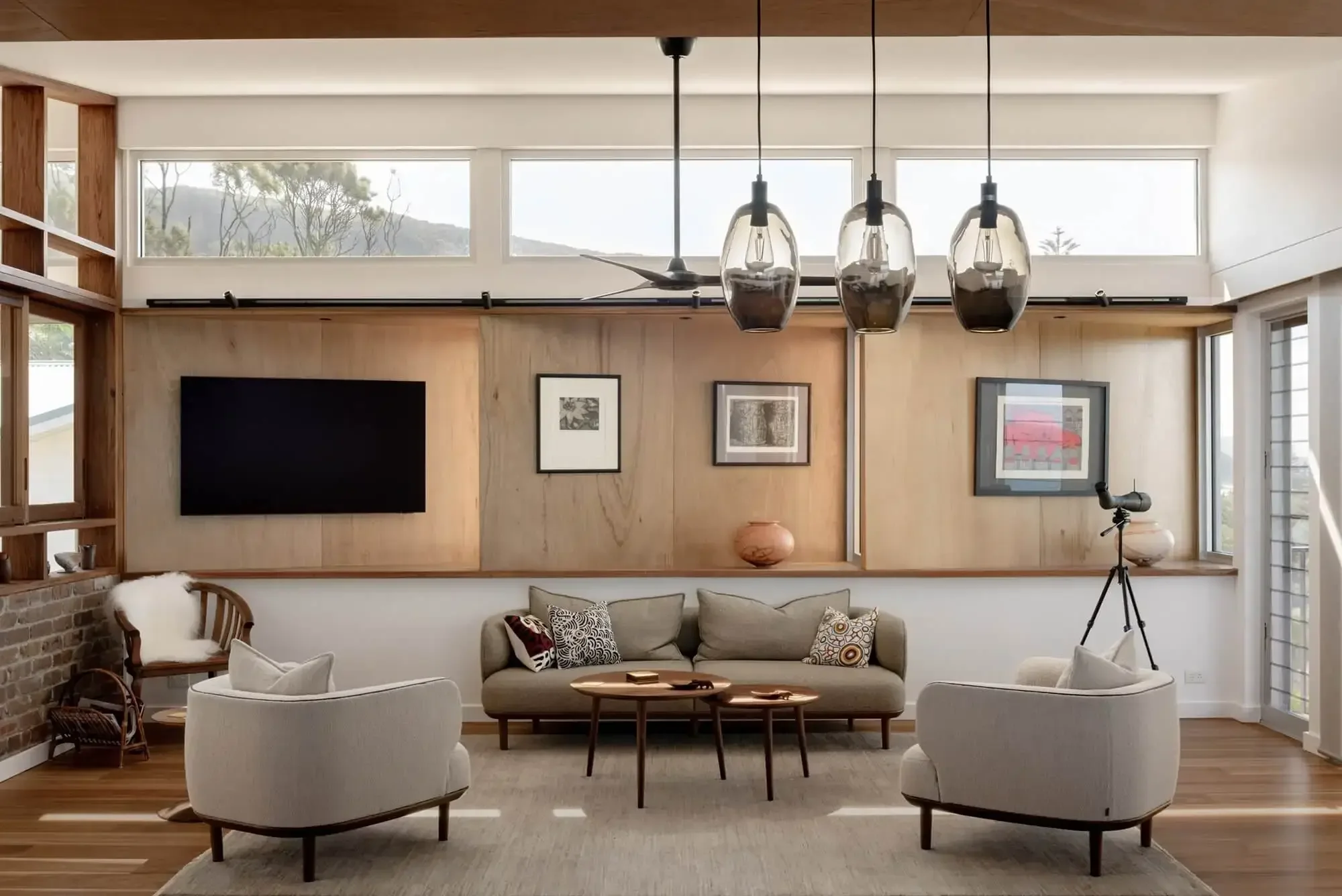 Modern living room with a neutral color palette, featuring a beige sofa, two armchairs, a flat-screen TV, framed art, a telescope, and pendant lighting. The room has large windows and wood accents.